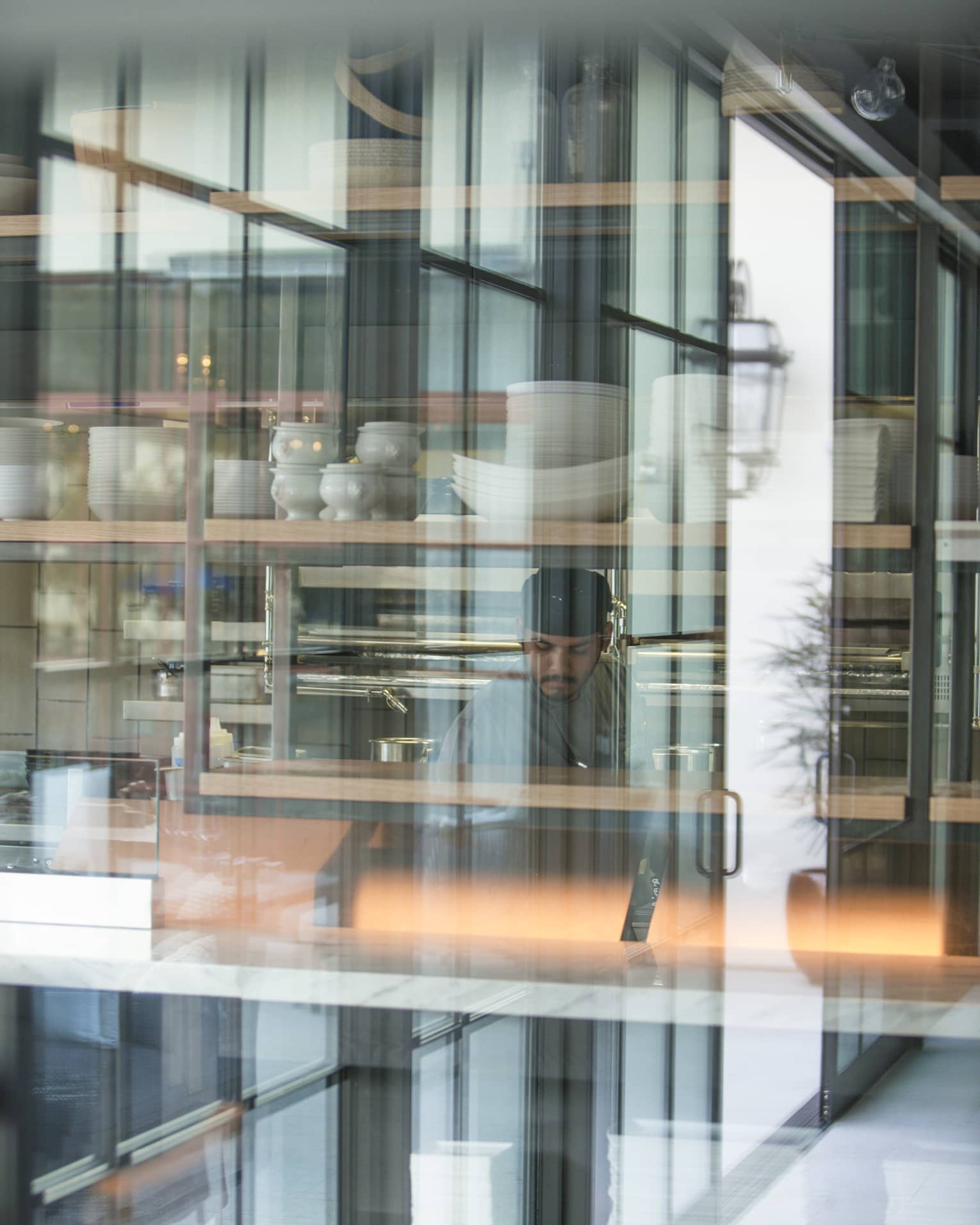 A chef working in a kitchen behind large glass windows, there are stacks of white dishes behind him.