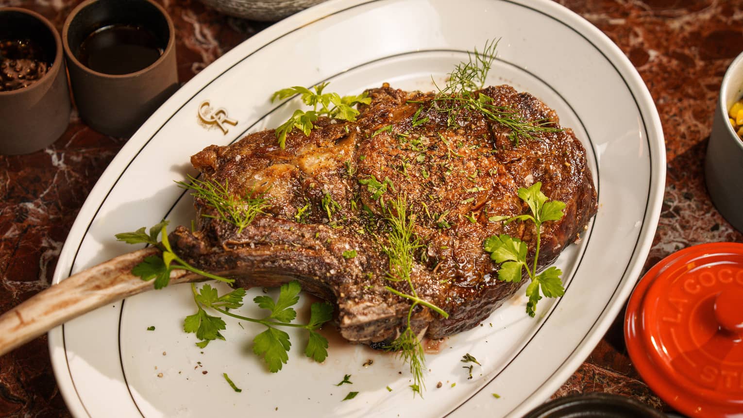 Black Angus Tomahawk garnished with sprigs of greenery and served on a white oval dish surrounded by various side dishes