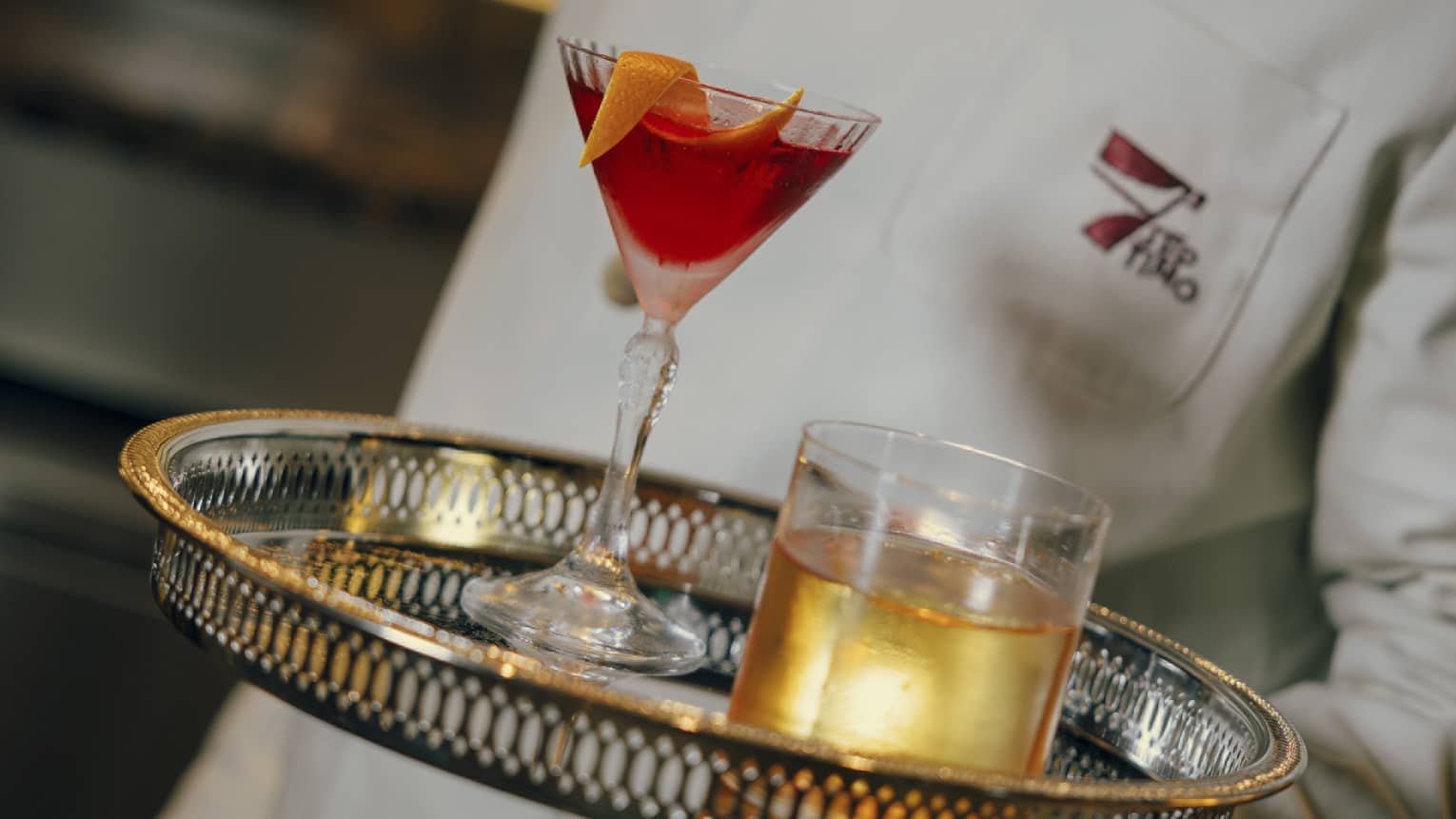Bartender carries two cocktails on a silver tray, one in a Martini glass and the other in a rocks glass