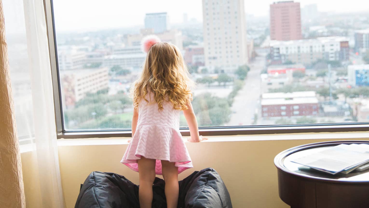 Young girl in pink dress and pink hair bow stands on bean bag chair, looks out window