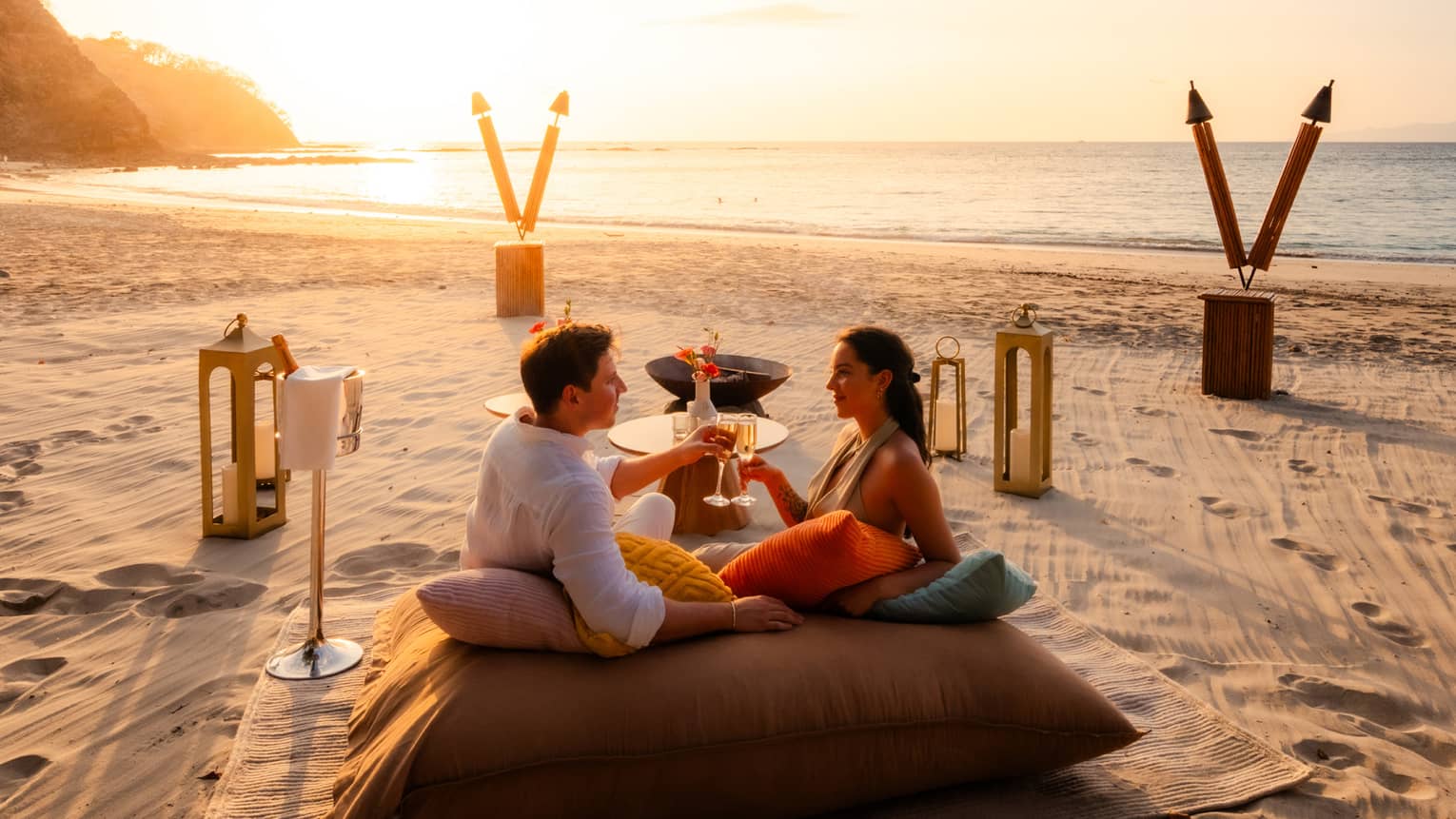 Couple sits on a large cushion on the beach with a private beach picnic set up around them