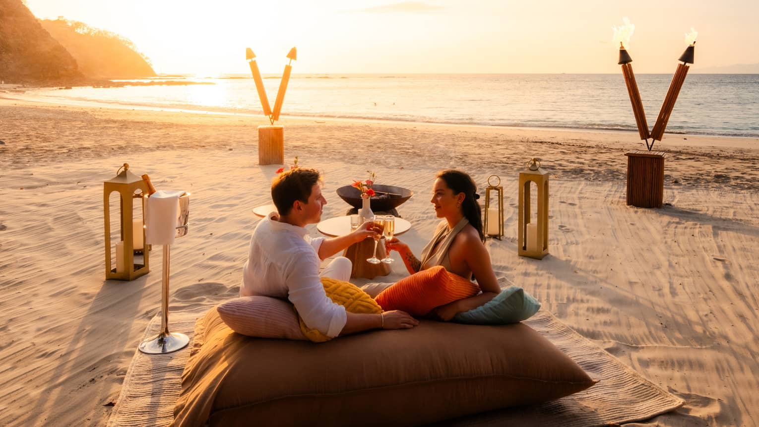 Couple sits together on a large cushion on the beach surrounded by a picnic set up just for two