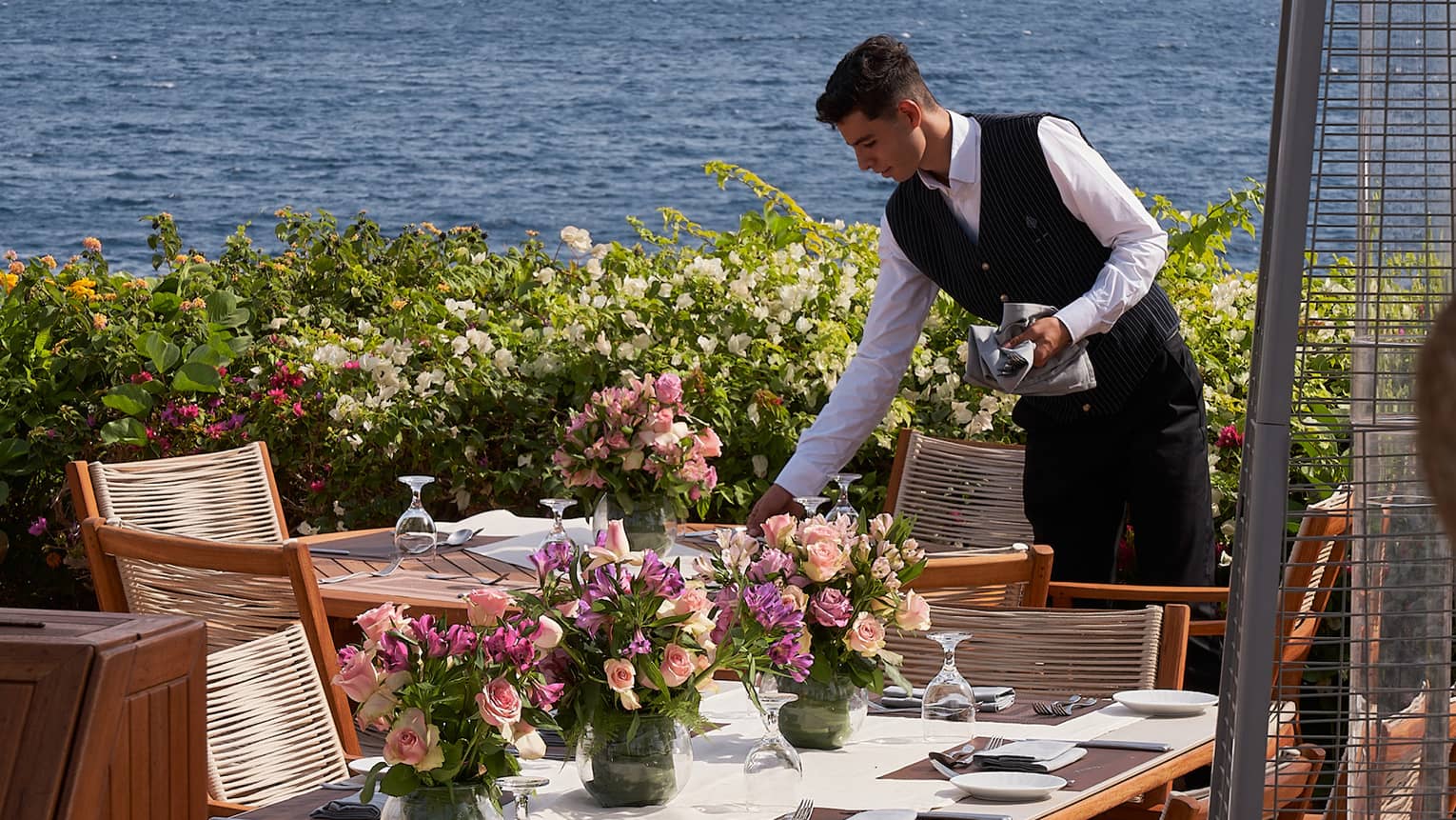 Waiter sets outdoor table with pink floral centrepieces, ocean and mountains in the background