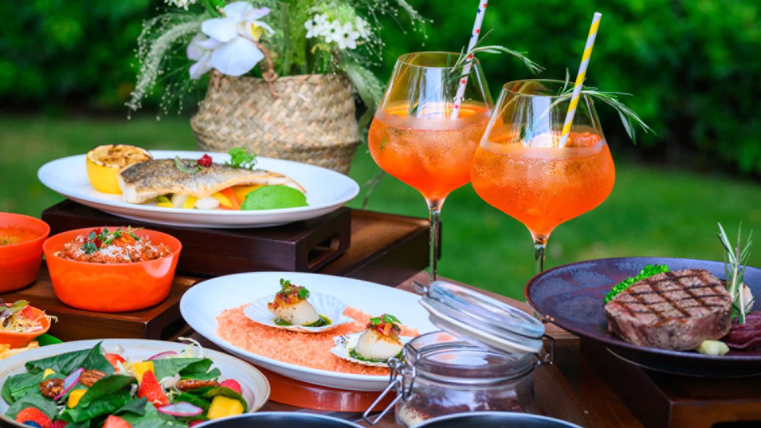 A spread of several courses of vibrant orange-themed fare with tropical drinks, aside a spray of delicate white orchids.