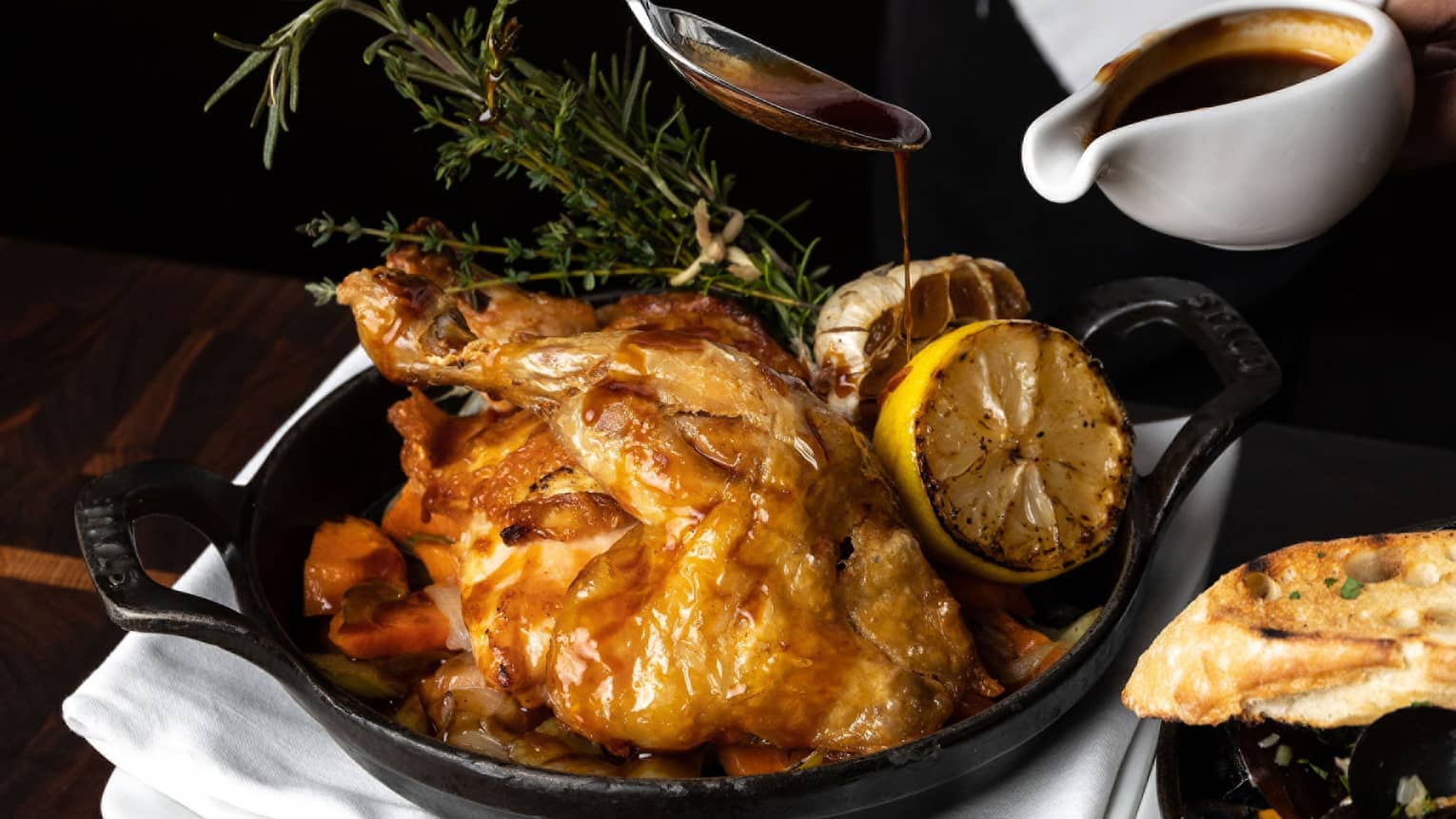 Chef's hands basting a full roasted chicken in an iron skillet, garnished with greenery and roasted lemons