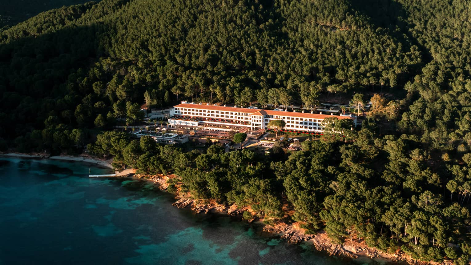An aerial view of a wooded, mountainous coastline with a resort building among the trees, blue water in the foreground, and mountain views and blue sky in the background