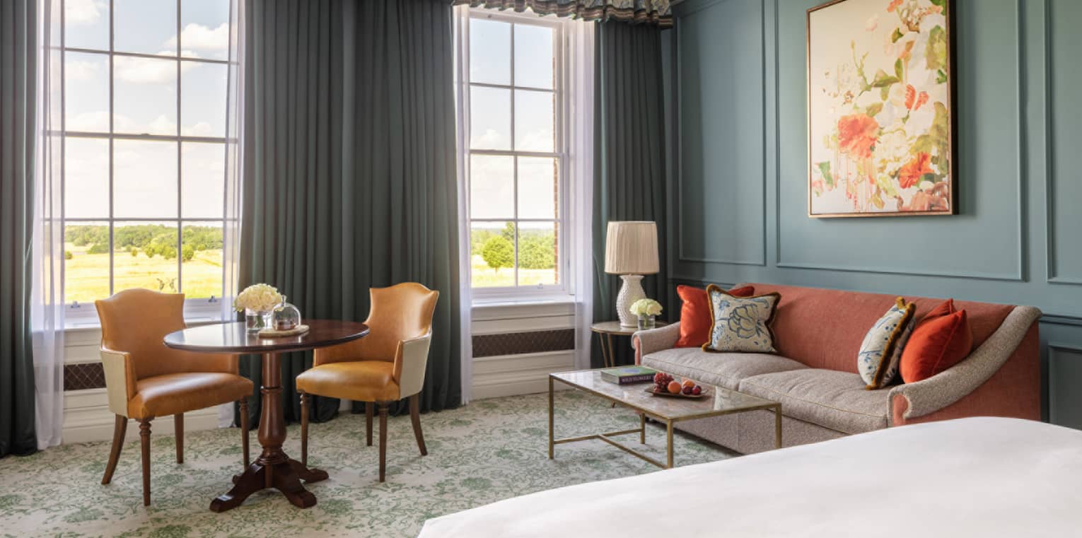 Guest room with bed, white linens, paisley carpeting, red and cream couch, small table for two, turquoise walls, twin windows with blue drapes