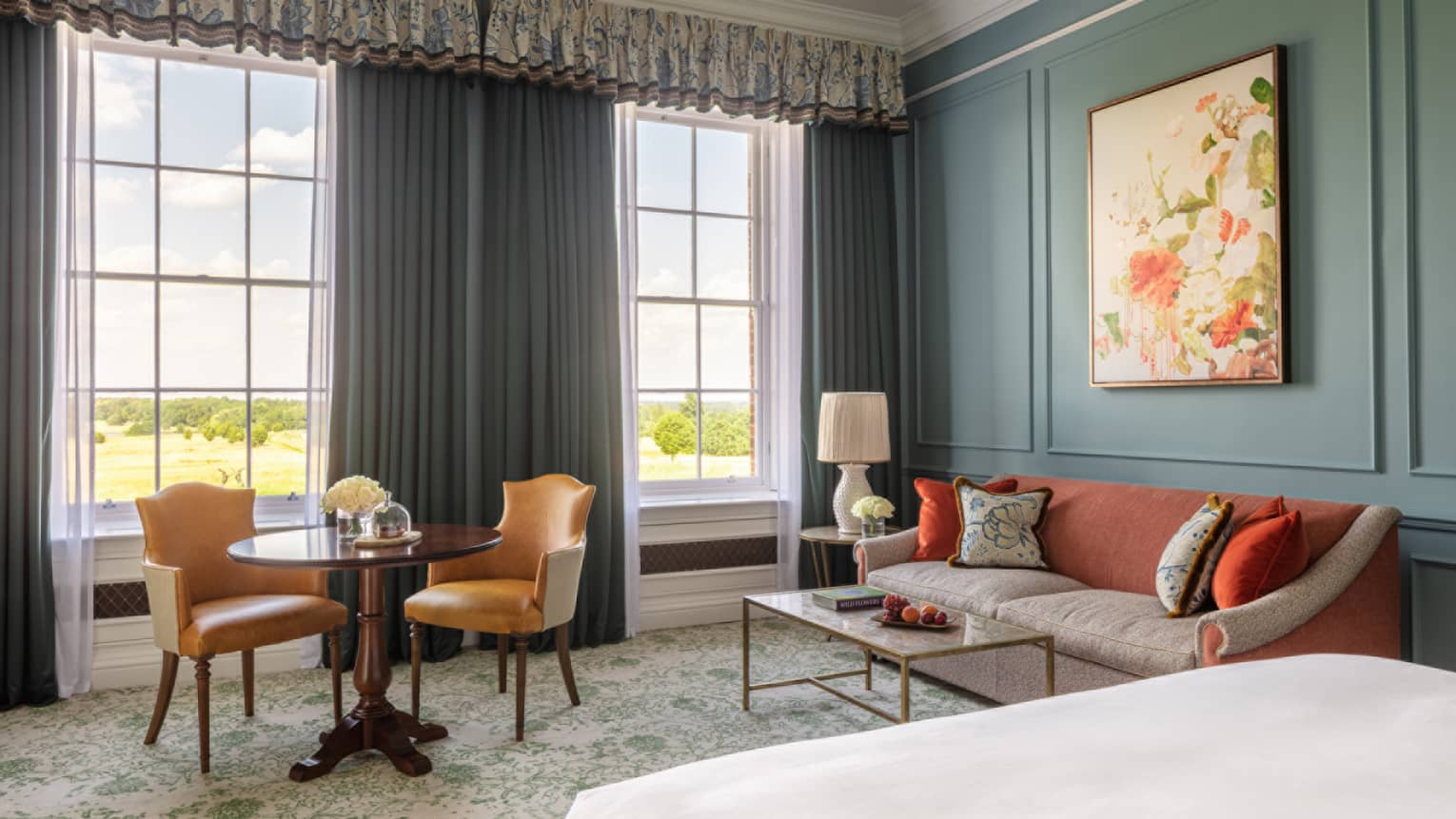 Guest room with bed, white linens, paisley carpeting, red and cream couch, small table for two, turquoise walls, twin windows with blue drapes