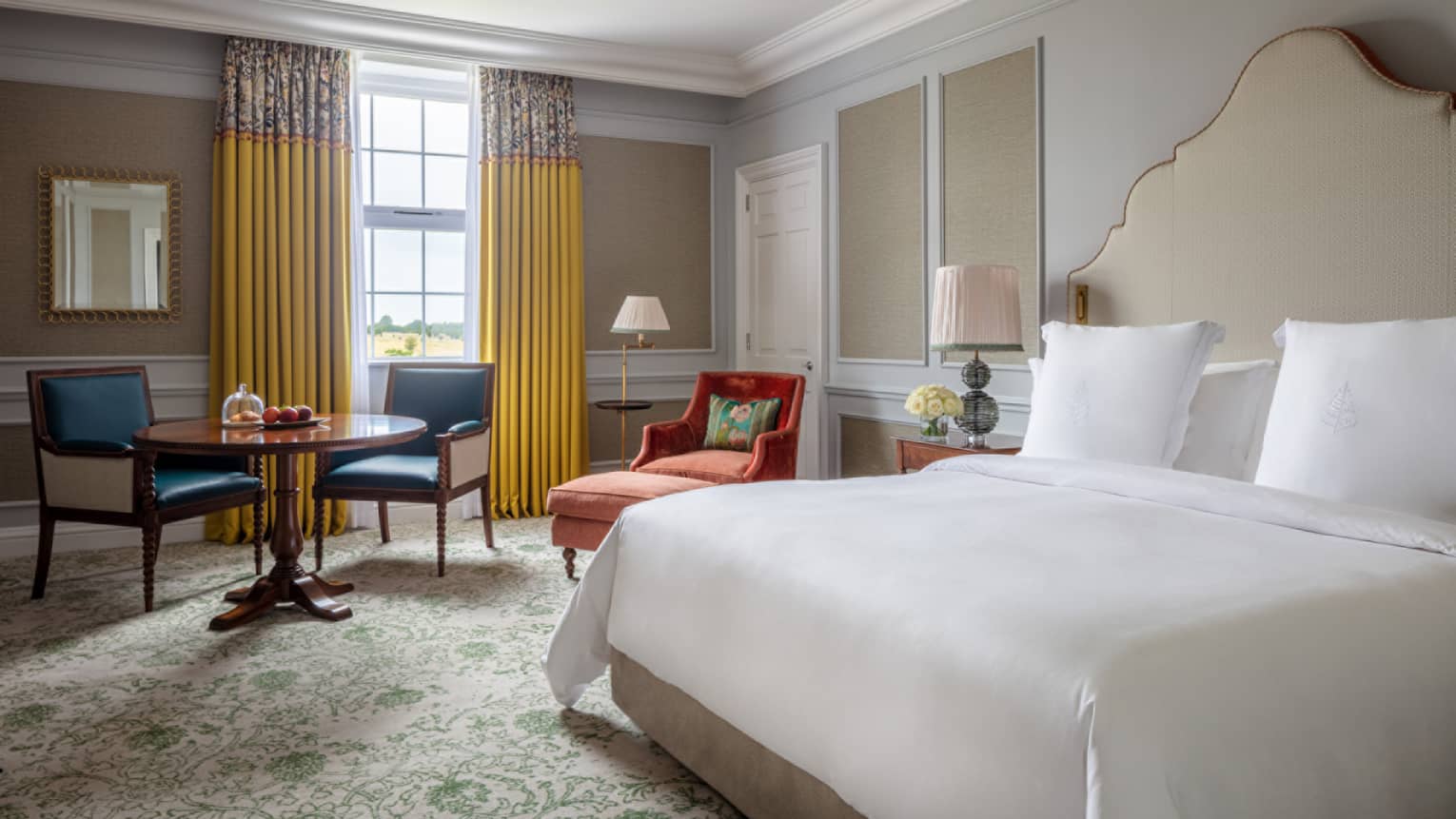 Guest room with king bed and white linens, red chaise, small round table and two chairs, window with yellow drapes