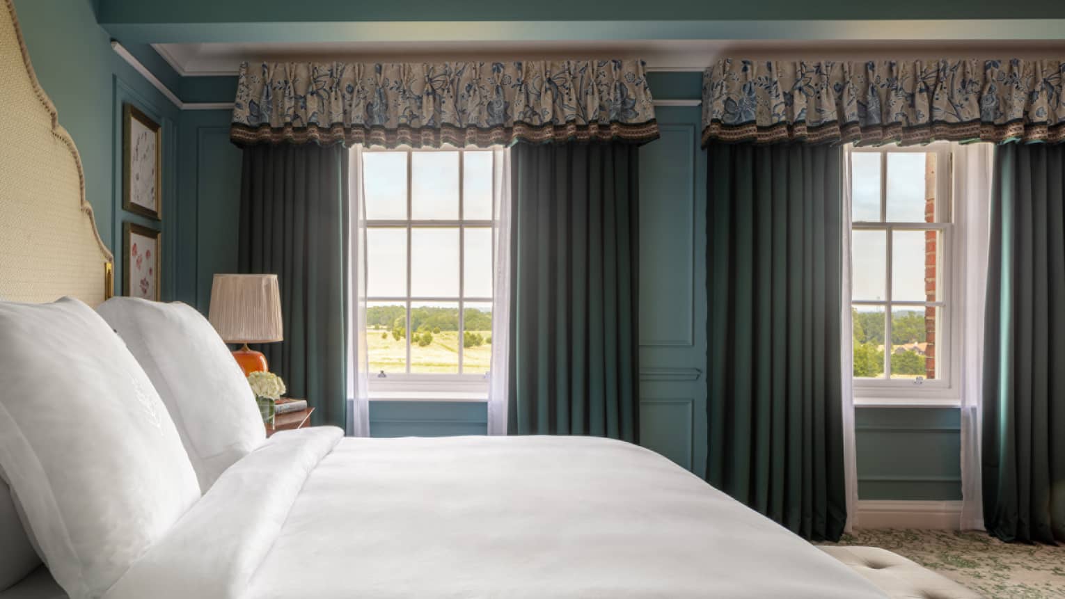 Guest room with bed with white linens, cream headboard and matching bench at foot of bed, twin windows with blue drapes