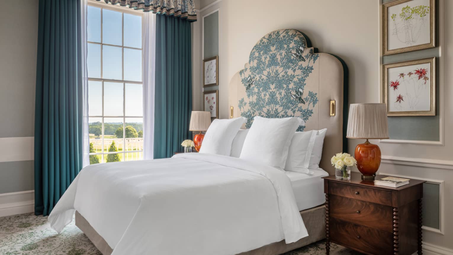 Guest room with Queen bed with white linens and embroidered headboard, cherry bedside table with orange lamp, floor-to-ceiling window with blue drapes