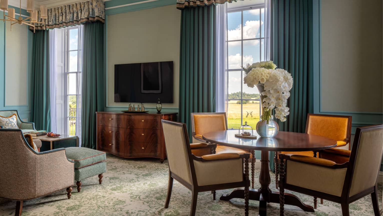Guest room with round wooden table and four dining chairs, cherry sideboard, flat screen TV, blue chaise, twin windows with blue drapes