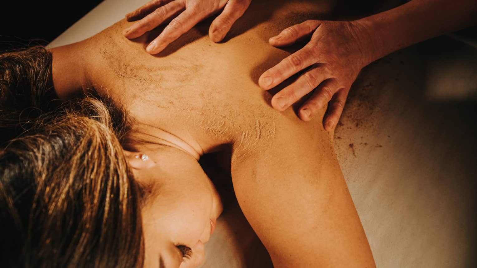 Two hands on woman's bare back as she lays on massage table in dimly lit room