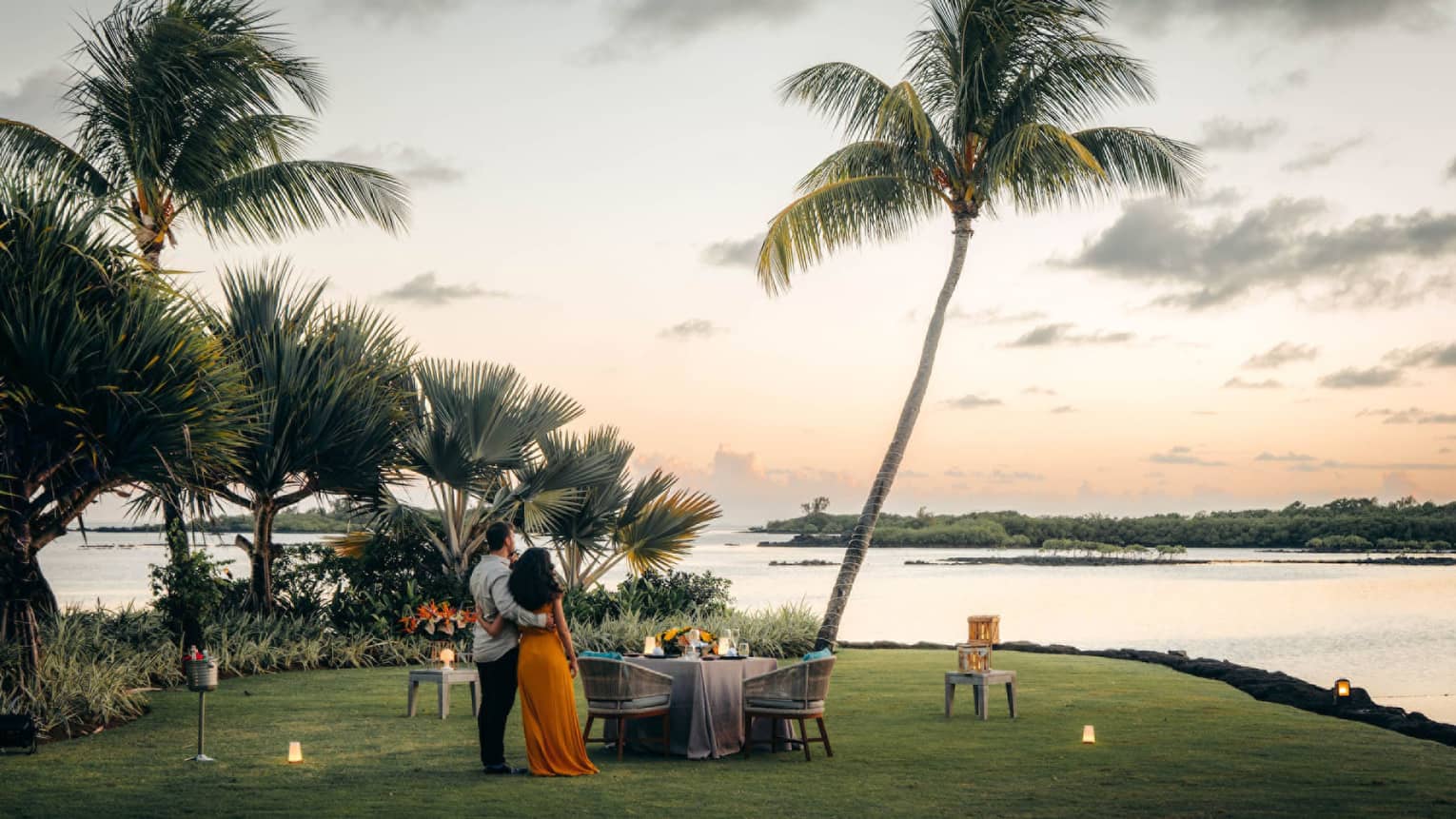 A couple stand on the shore, arms around each other, gazing toward the ocean; set table with two chairs beside swaying palms.