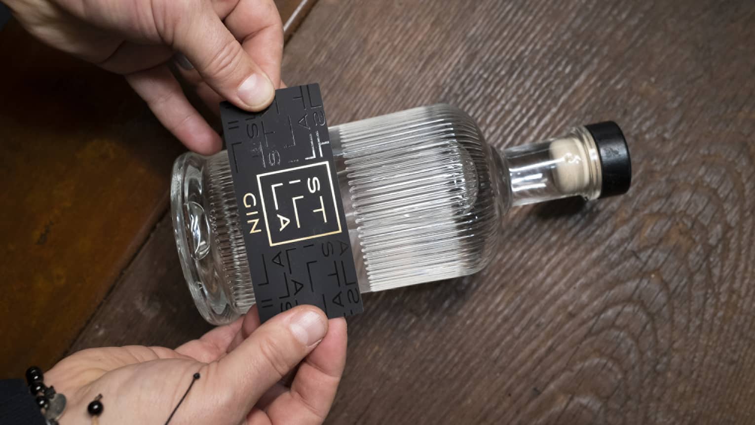 Hands placing black Stilla Gin label on white beveled glass bottle laying on wooden table