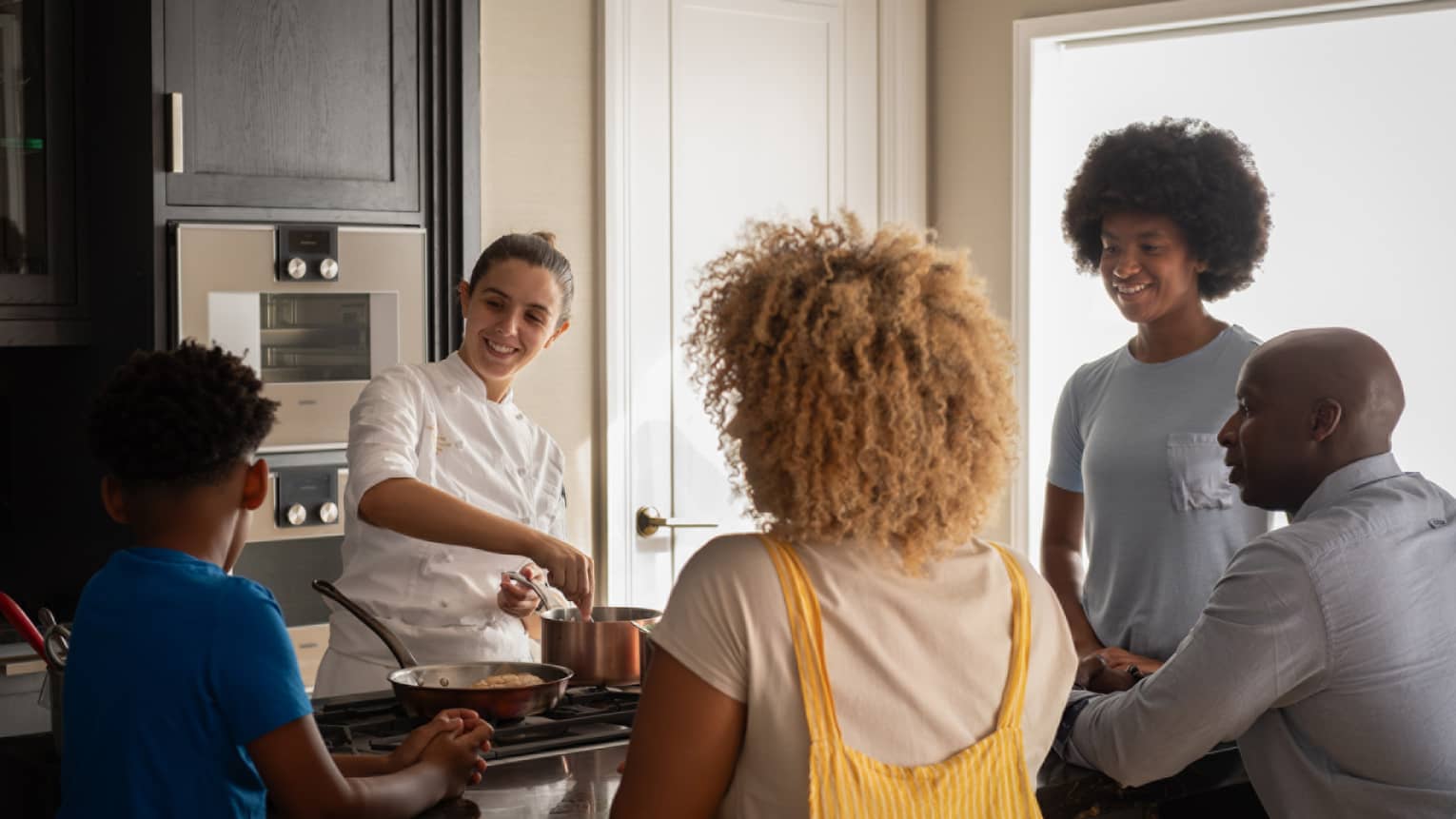 A family of four gathers around a kitchen-island cooktop watching a smiling chef prepare food in copper cookware. 