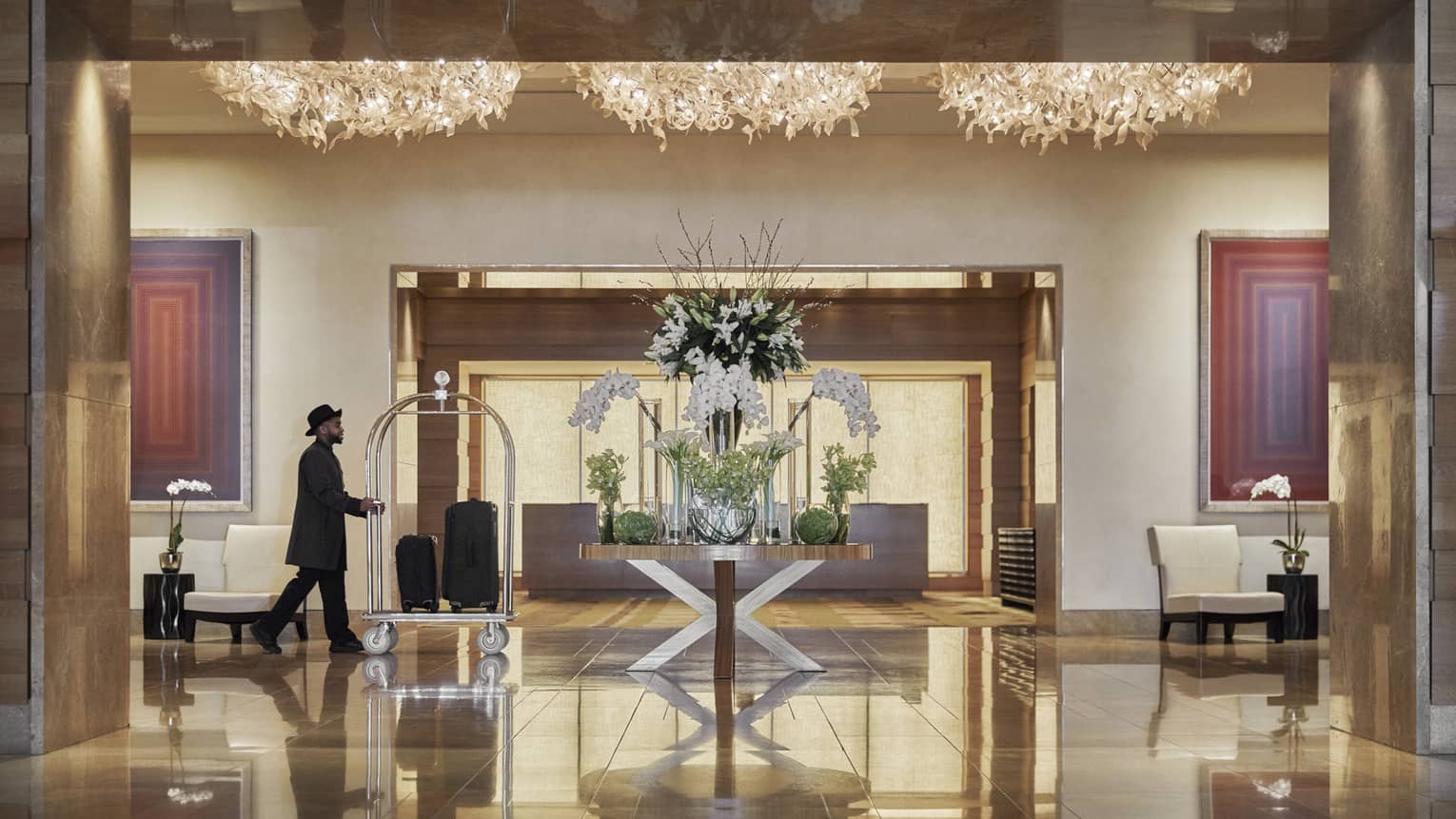 A man in black clothes pushing a trolly of suitcases across a hotel lobby that is lit by gold chandaliers.