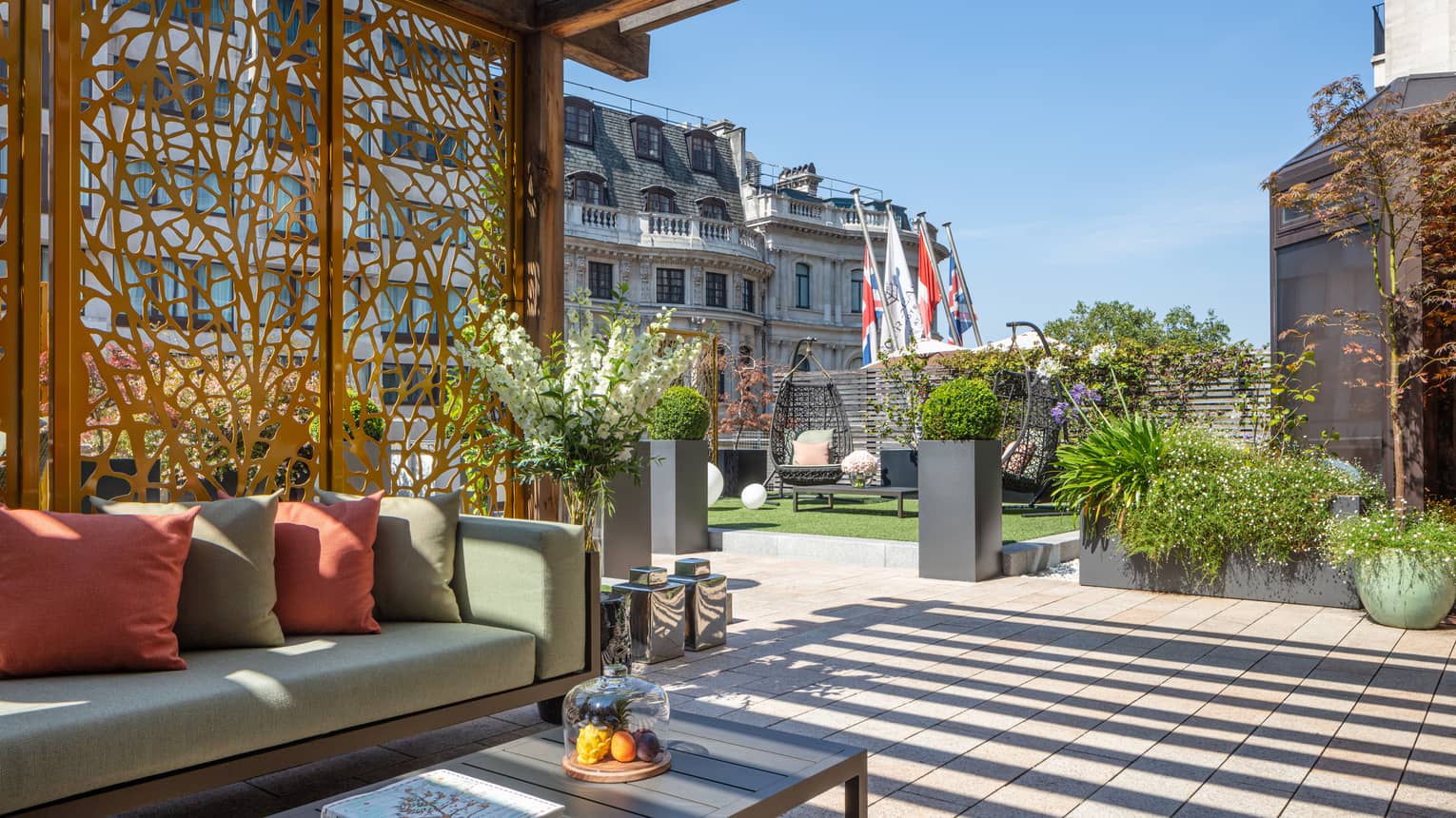 The Terrace Suite at Four Seasons London Park Lane in Mayfair features an expansive and beautifully designed outdoor terrace. The space is adorned with modern outdoor furniture including a sleek sofa with plush cushions and a low coffee table, perfect for relaxation or entertaining. The terrace is surrounded by lush potted plants, sculptural elements, and features decking that adds to the sophisticated ambiance. The view includes the tops of surrounding historic London buildings, creating a luxurious and serene urban garden atmosphere.