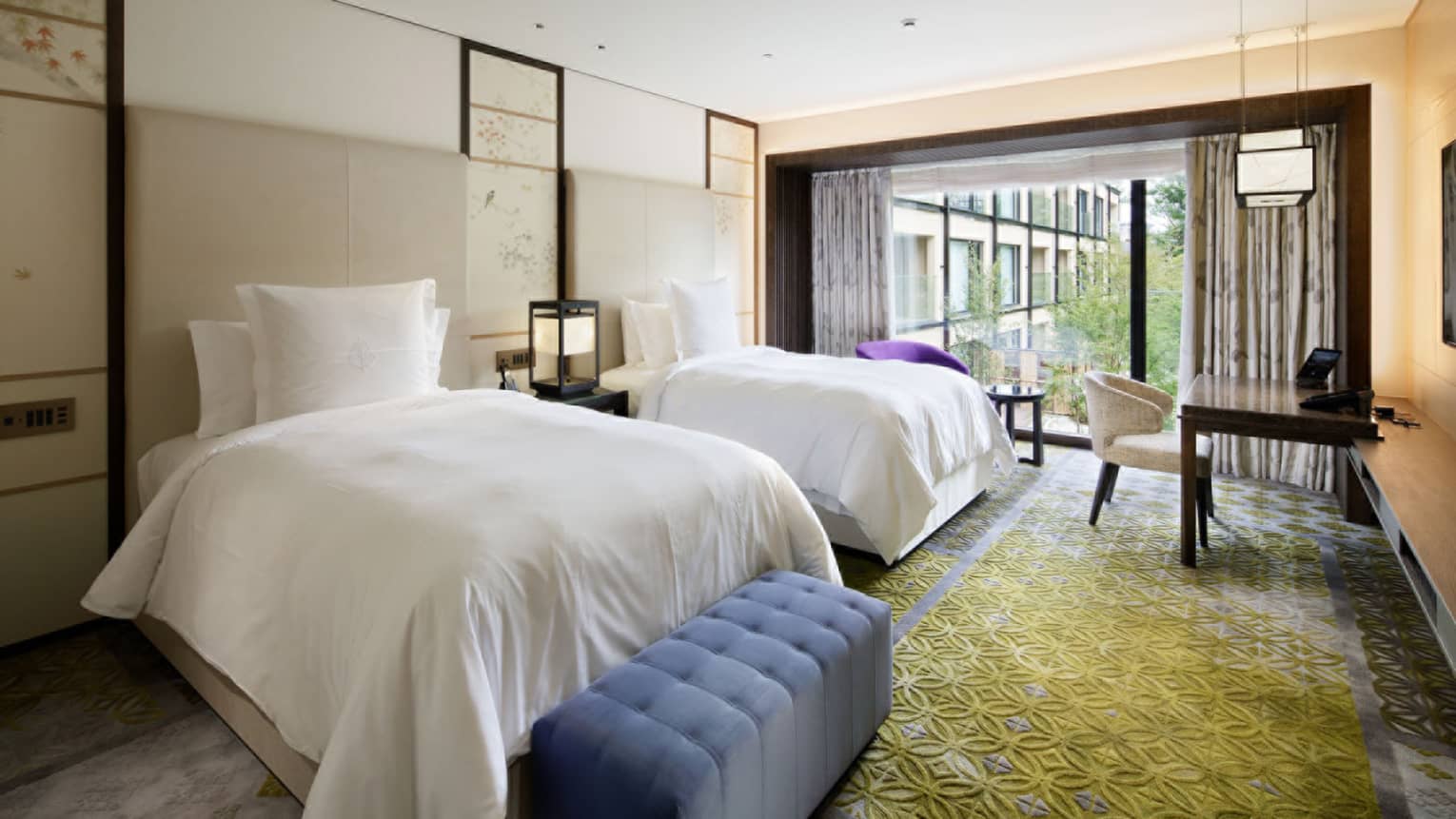 Bright hotel room with two beds, a soft blue tufted bench at the foot of the bed, a work desk and floor-to-ceiling windows overlooking greenery
