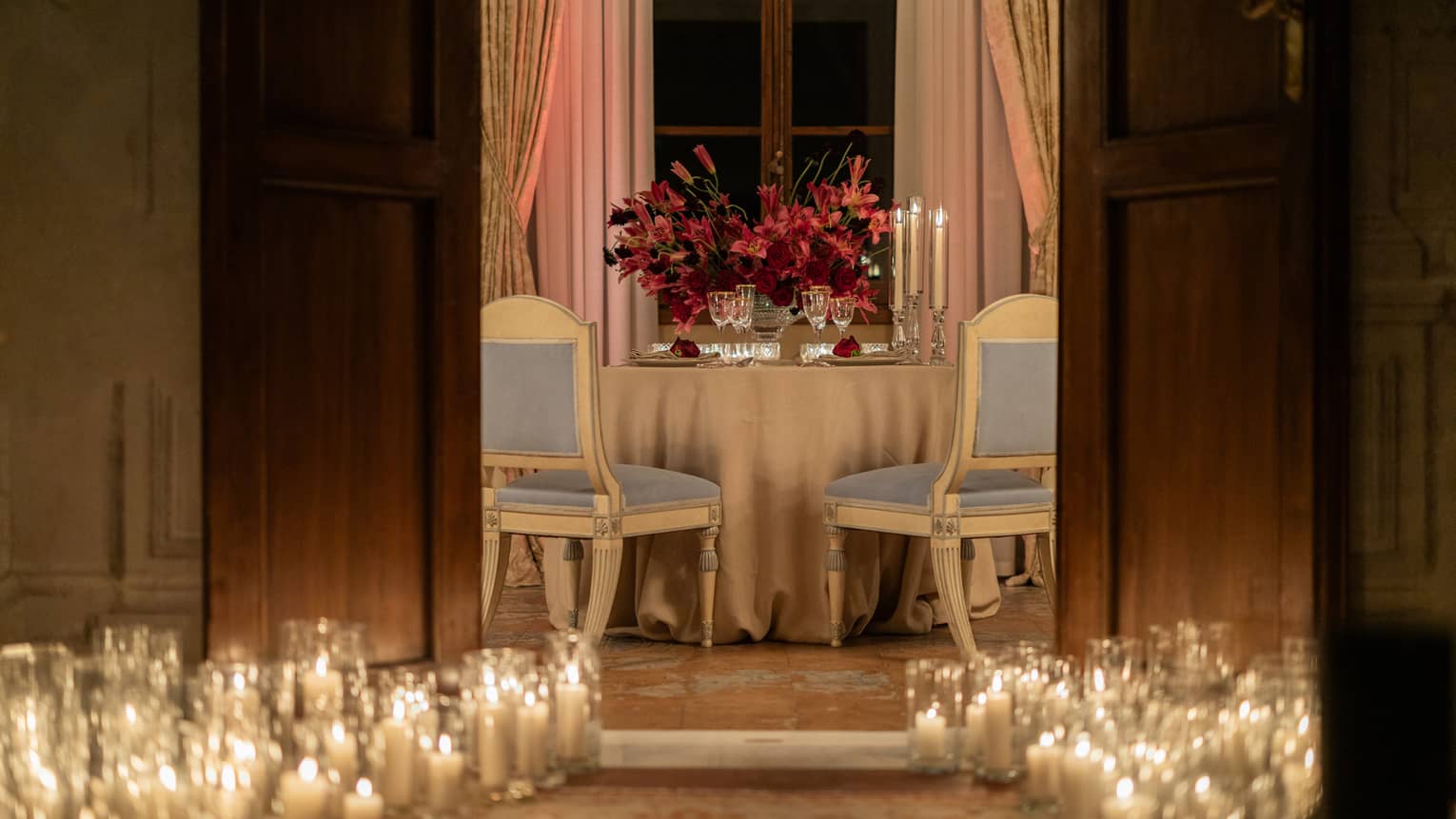 Lit hurricanes lining an entrance to a private meeting space, where a round table set for two toped with a large flower arrangement awaits