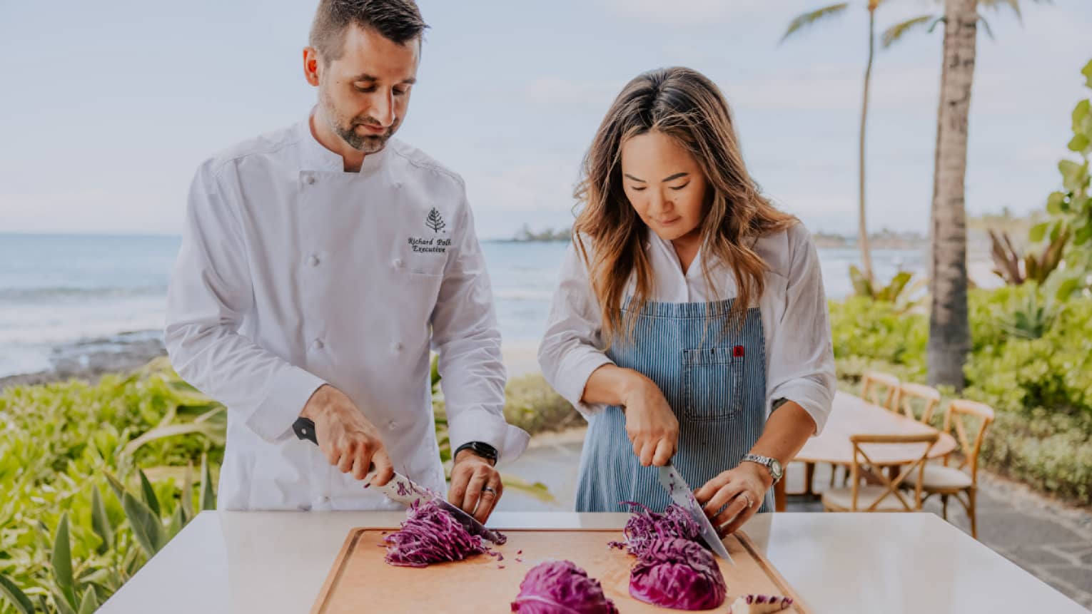 A woman and a man in a chef's jacket practice thinly slicing vegetables