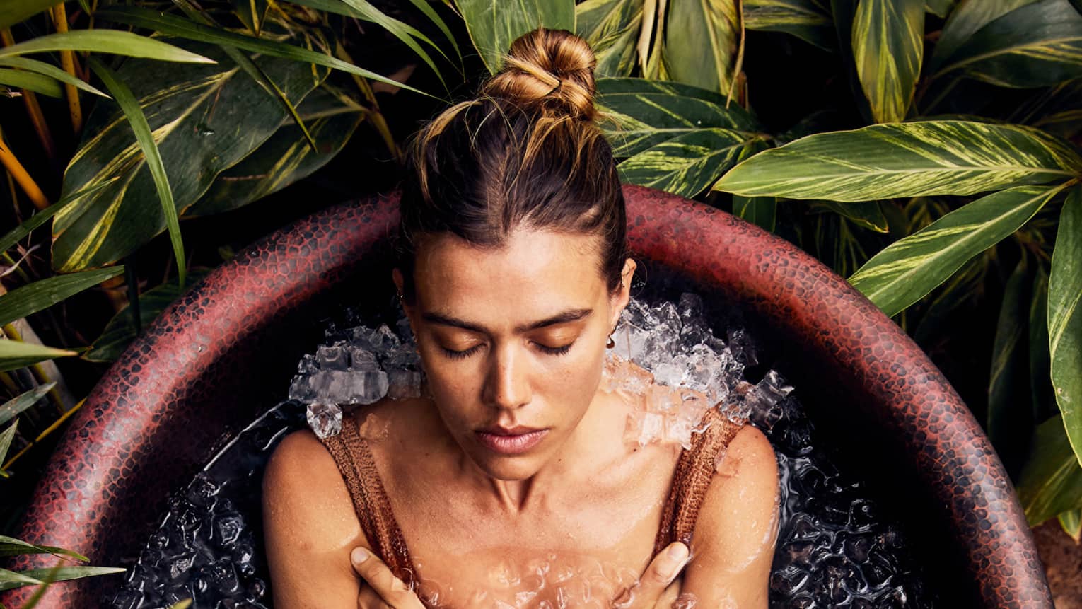 A guest, wearing a bathing suit, is sitting and relaxing in a rustic bathtub full of ice with foliage encircling the tub.