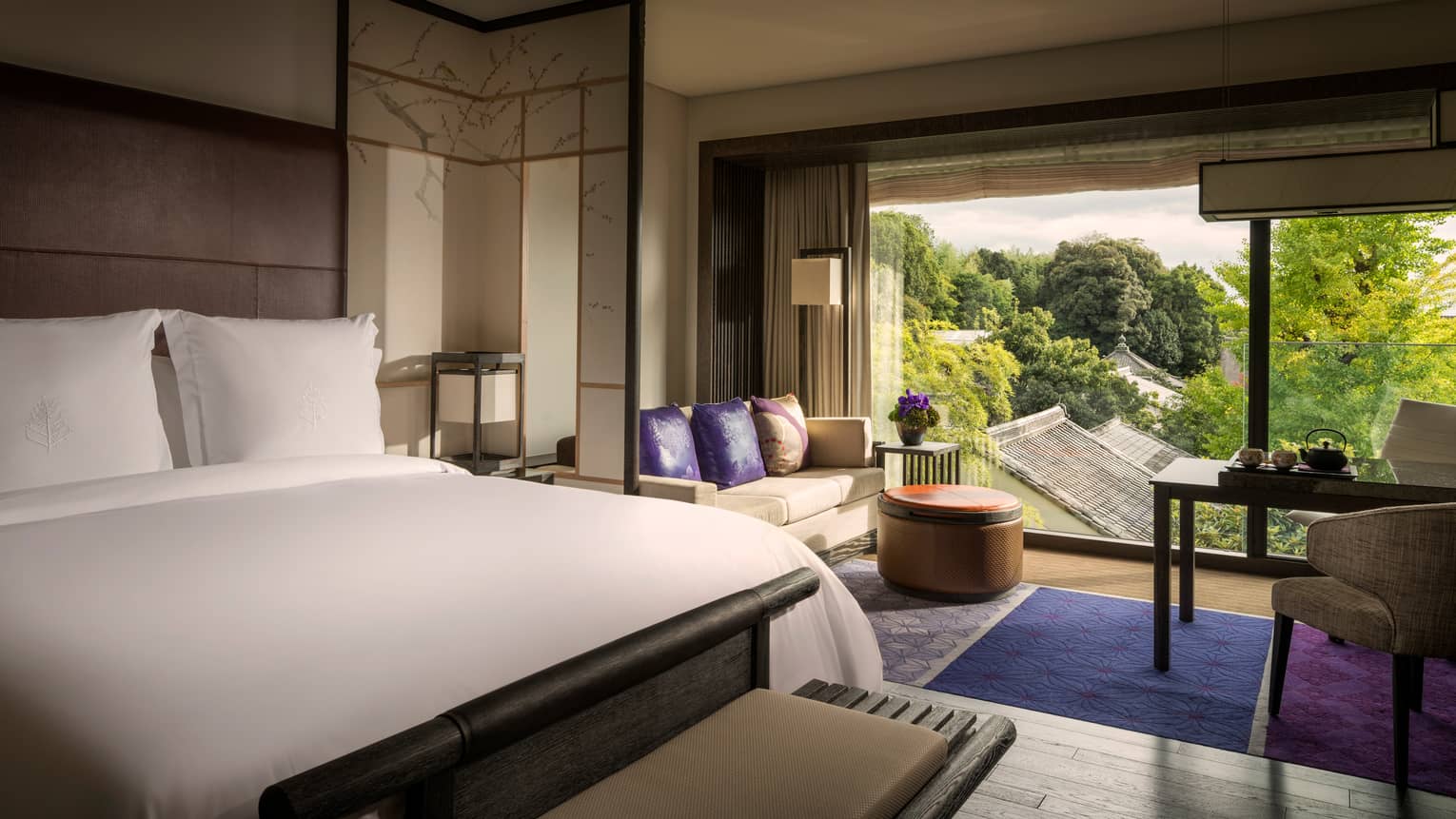 Hotel bed, loveseat with purple accent pillows, purple rug, floor-to-ceiling window