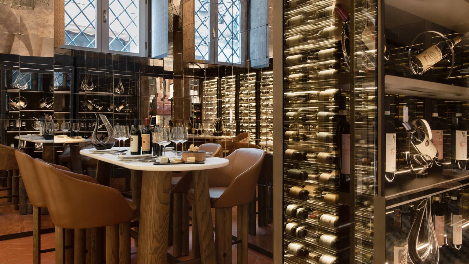 A table for four set within the mirrored walls of a wine cellar stocked with bottles displayed horizontally in glass cases.