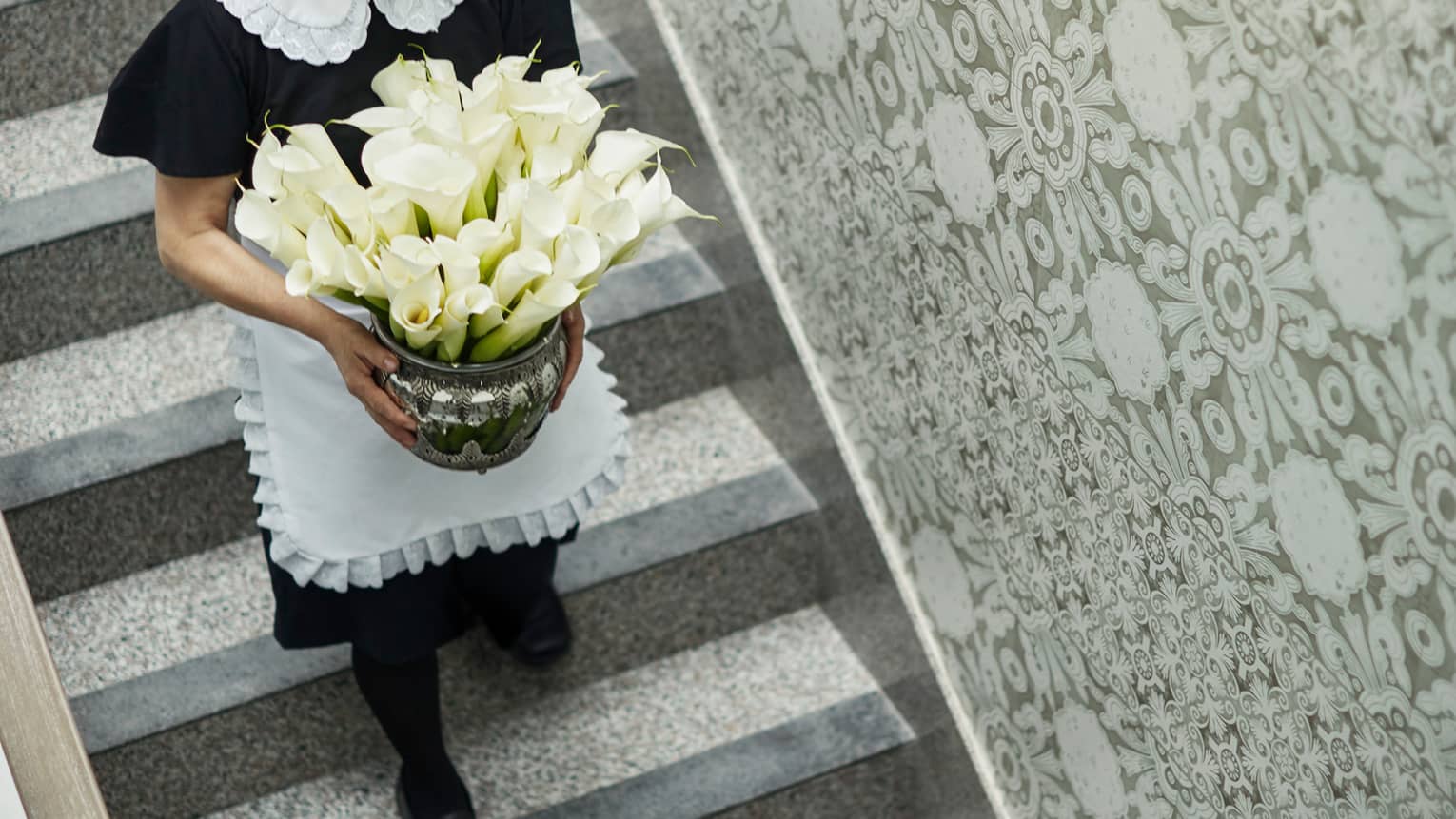 Housekeeper in black dress and white frilly apron walks down staircase holding vase with white flowers