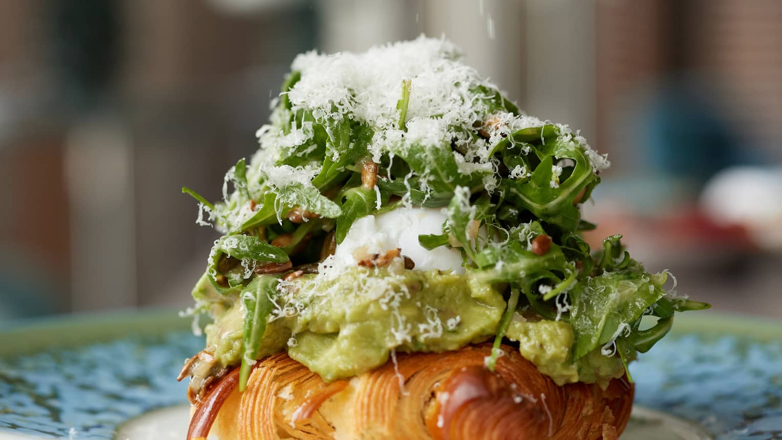 A close-up of a plate holding a croissant covered with avocado, egg and arugula salad topped with grated parmesan. 