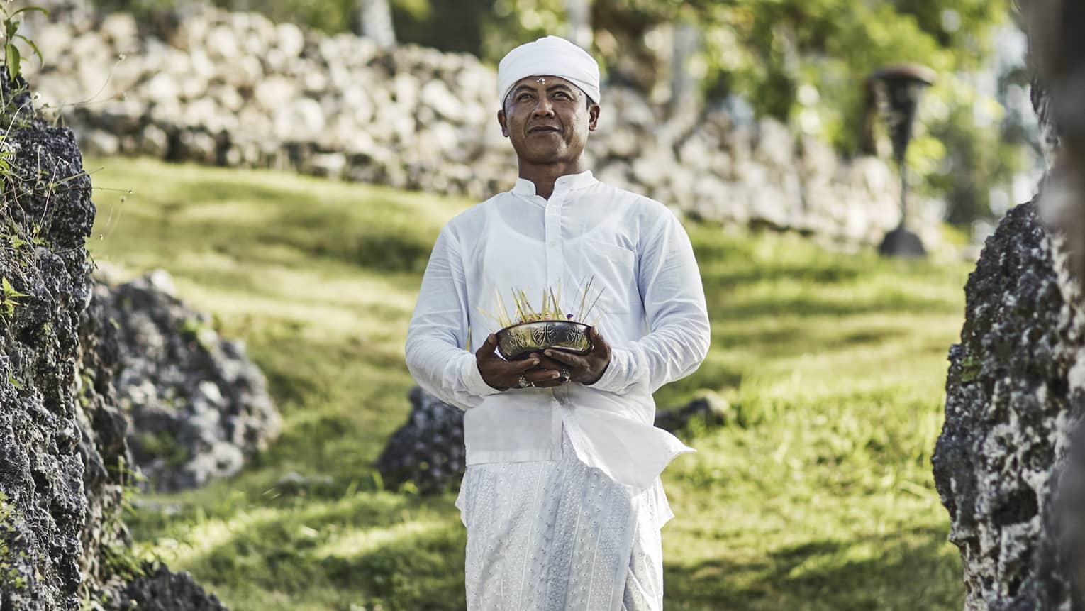 A priest in white attire stands on a stepping stone on a grassy hill, eyes gazing ahead, and holds a bowl of incense sticks.