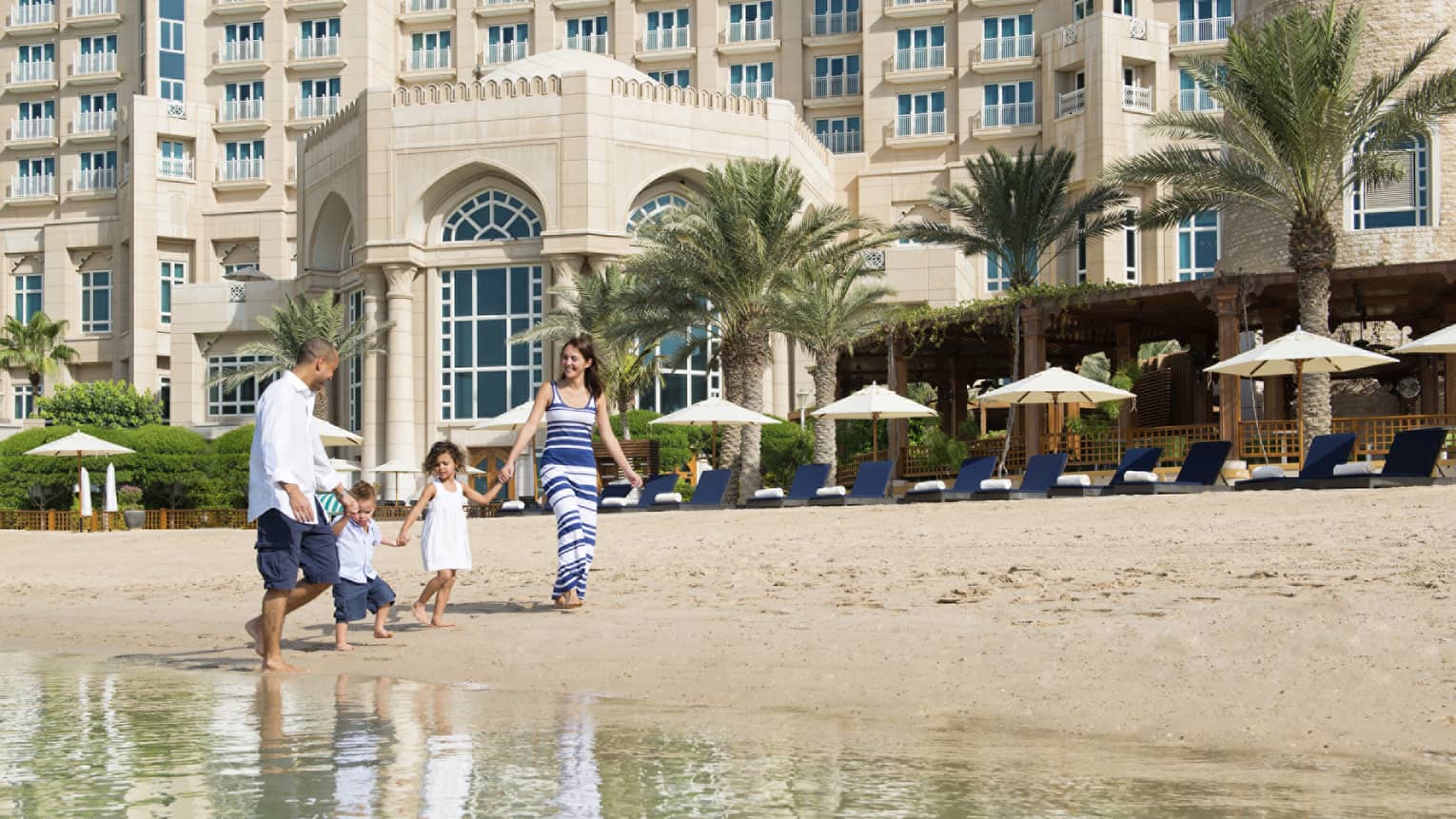 Woman and man hold hands of two young children, walk barefoot in sand along beachfront