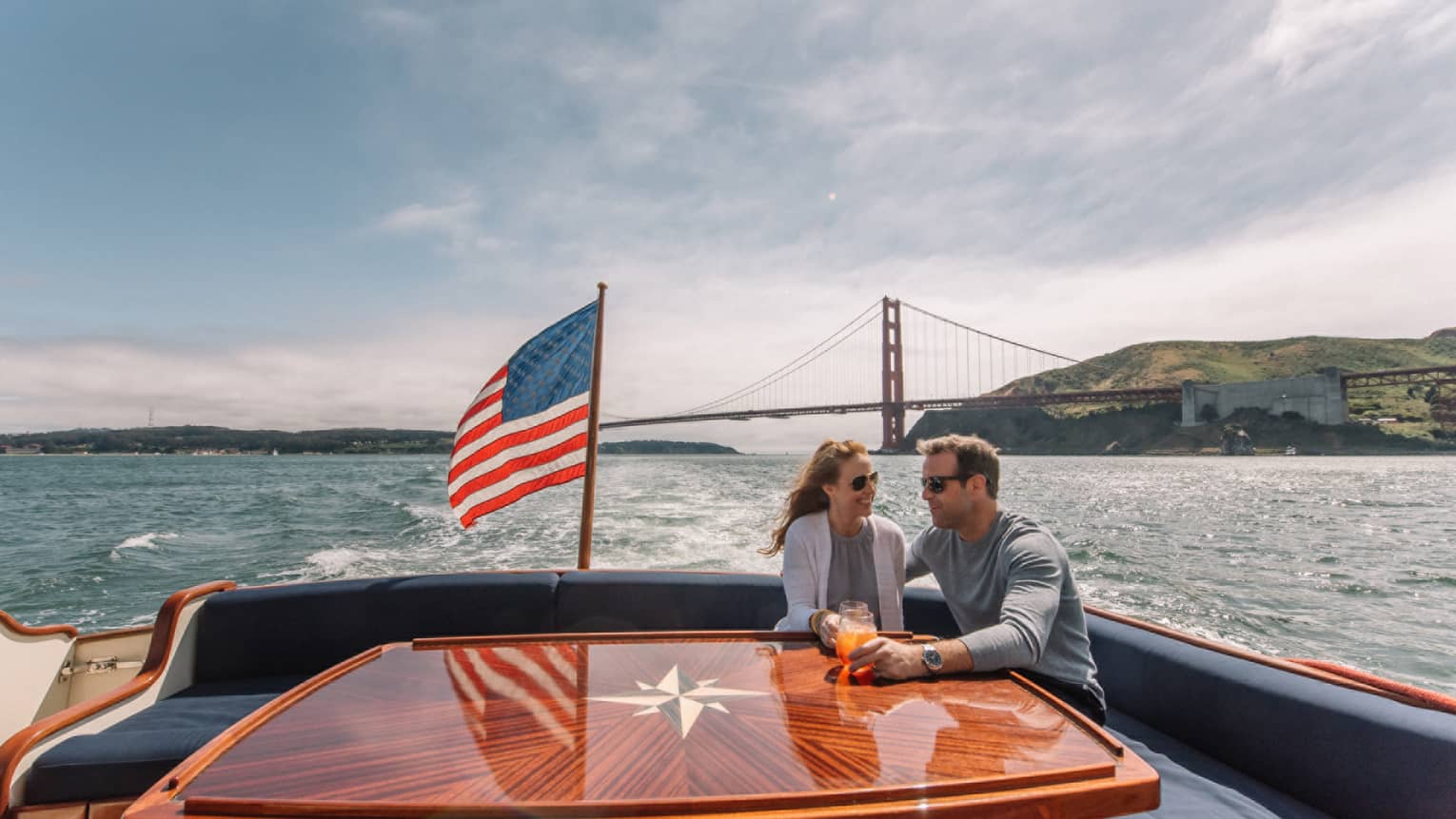 A man and woman on the back of a boat with a metal bridge in the distance behind them and an American flag on the boat.