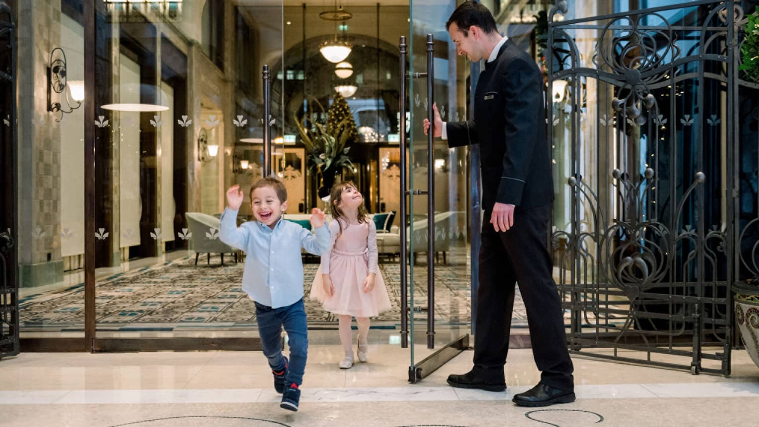 A young boy and girl walking through a hotel door being held open by a man in a suit.