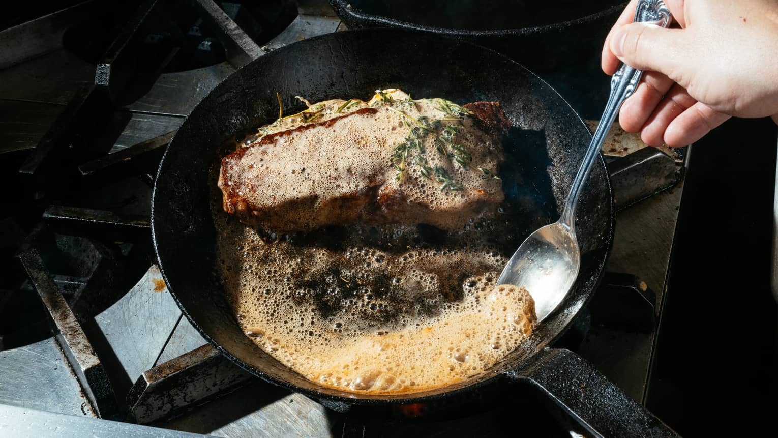 A skillet with a garnished cooked cut of meat and a pool of bubbling sauce, which a chef is scooping with a spoon