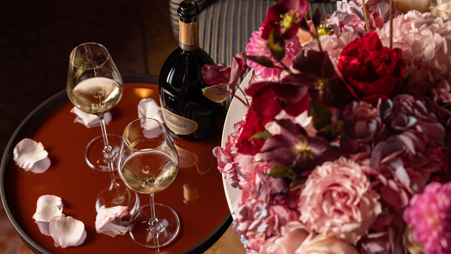 Two glasses of white wine on a brown table, surrounded by scattered rose petals and placed next to a large bouquet of flowers.