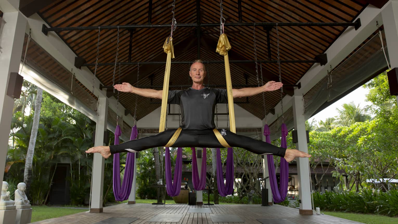 In a covered gazebo, the instructor smiles serenely while suspended in a gold silk sling, arms and legs outstretched..