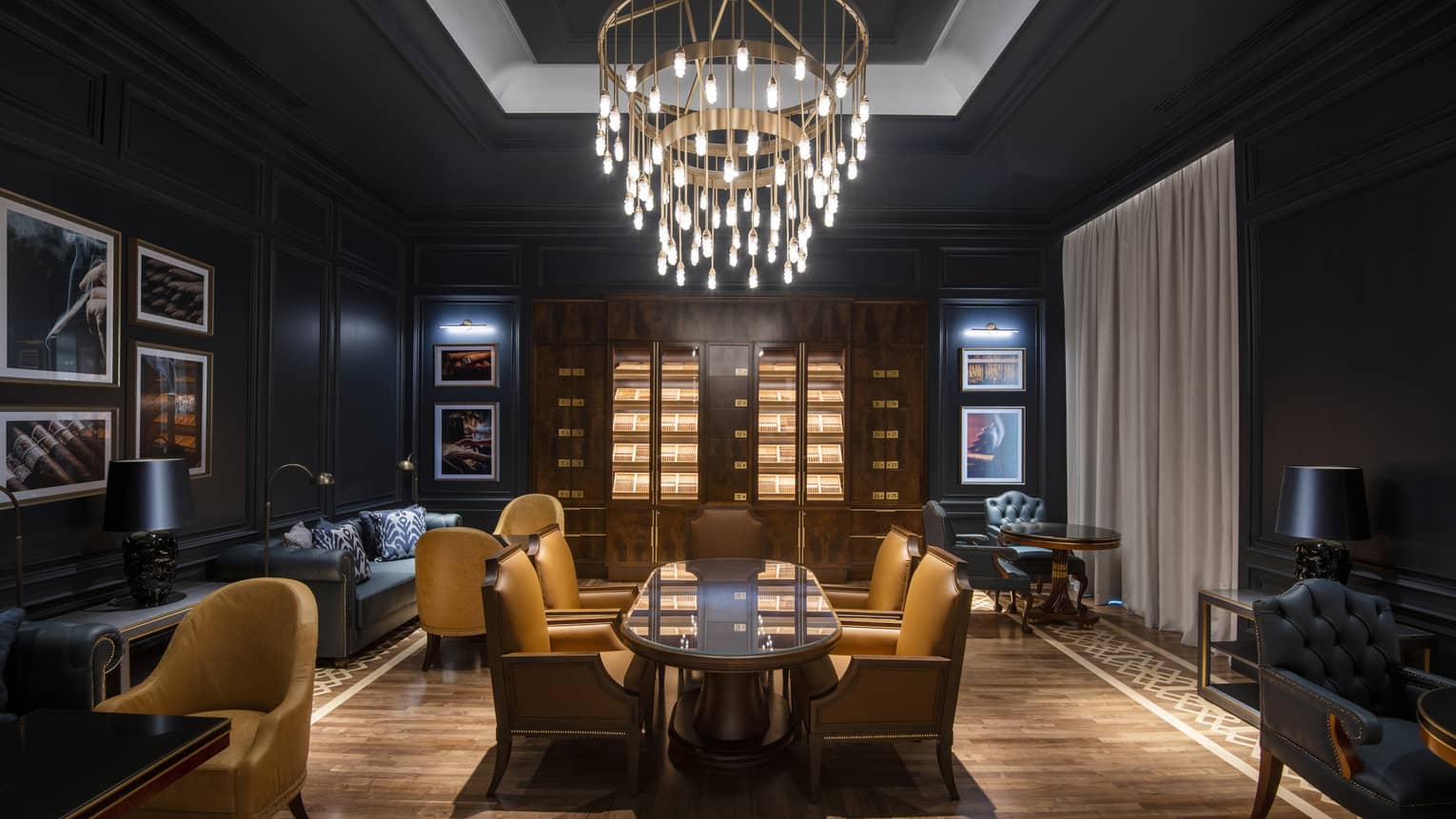 A lounge area with long wood table, dark sofas and walls and a hanging chandelier.