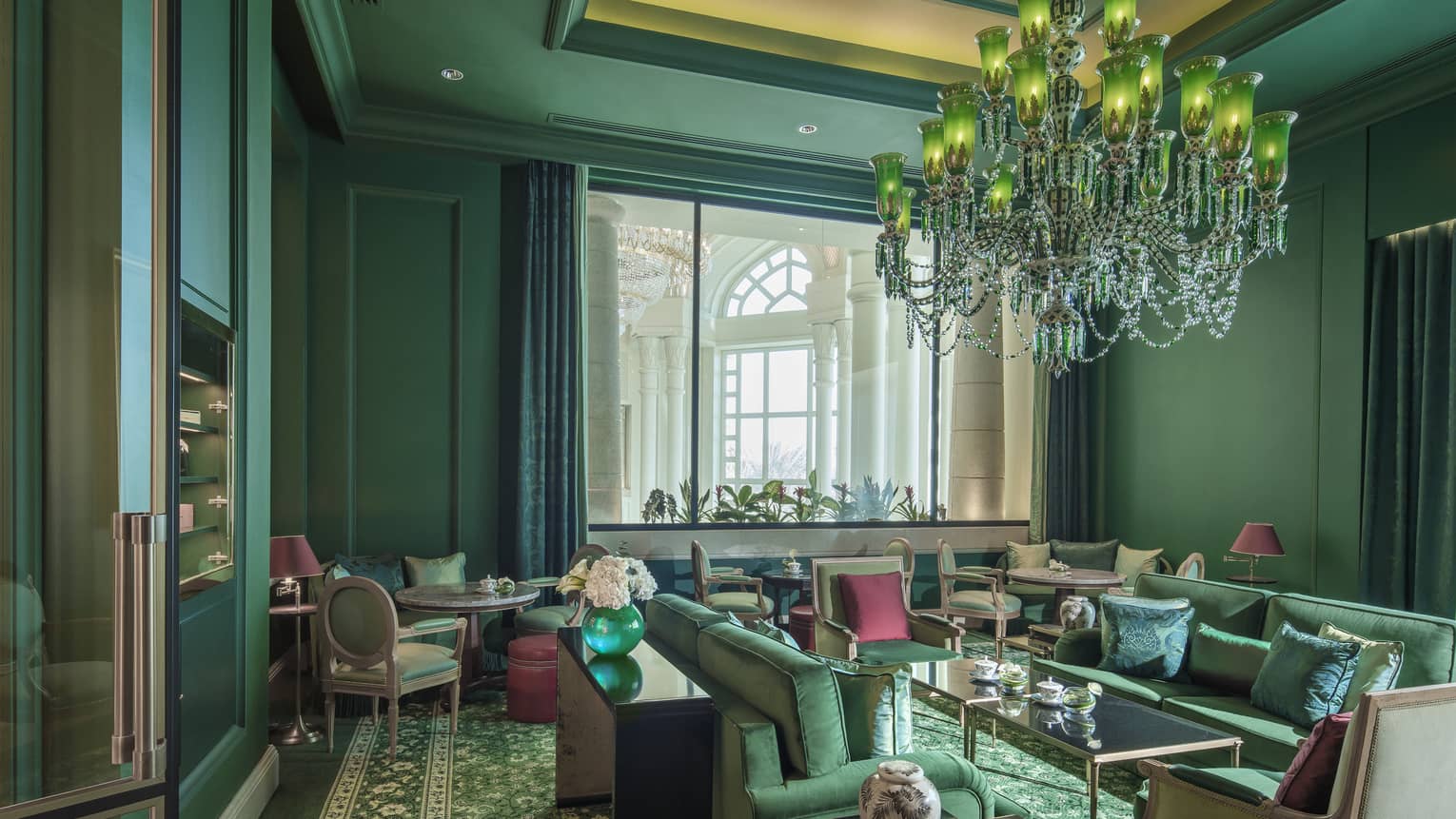 A forest green sitting room with over a dozen velveteen chairs, and a crystal chandelier. 