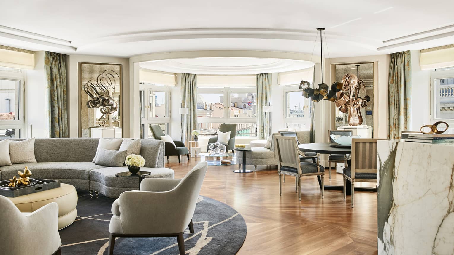 Spacious luxury suite with curved grey sectional, plush armchairs, abstract sculptures, marble accents, modern dining area and large windows with city views