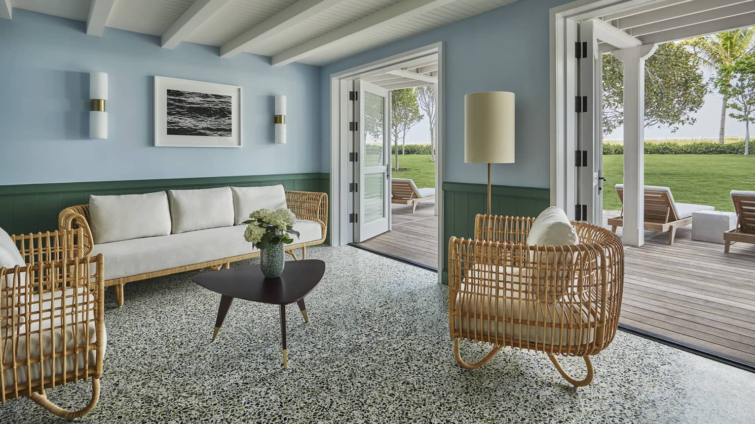 White-cushioned rattan furniture in a spacious living room of a private cabana, with double doors opening onto a scenic deck.