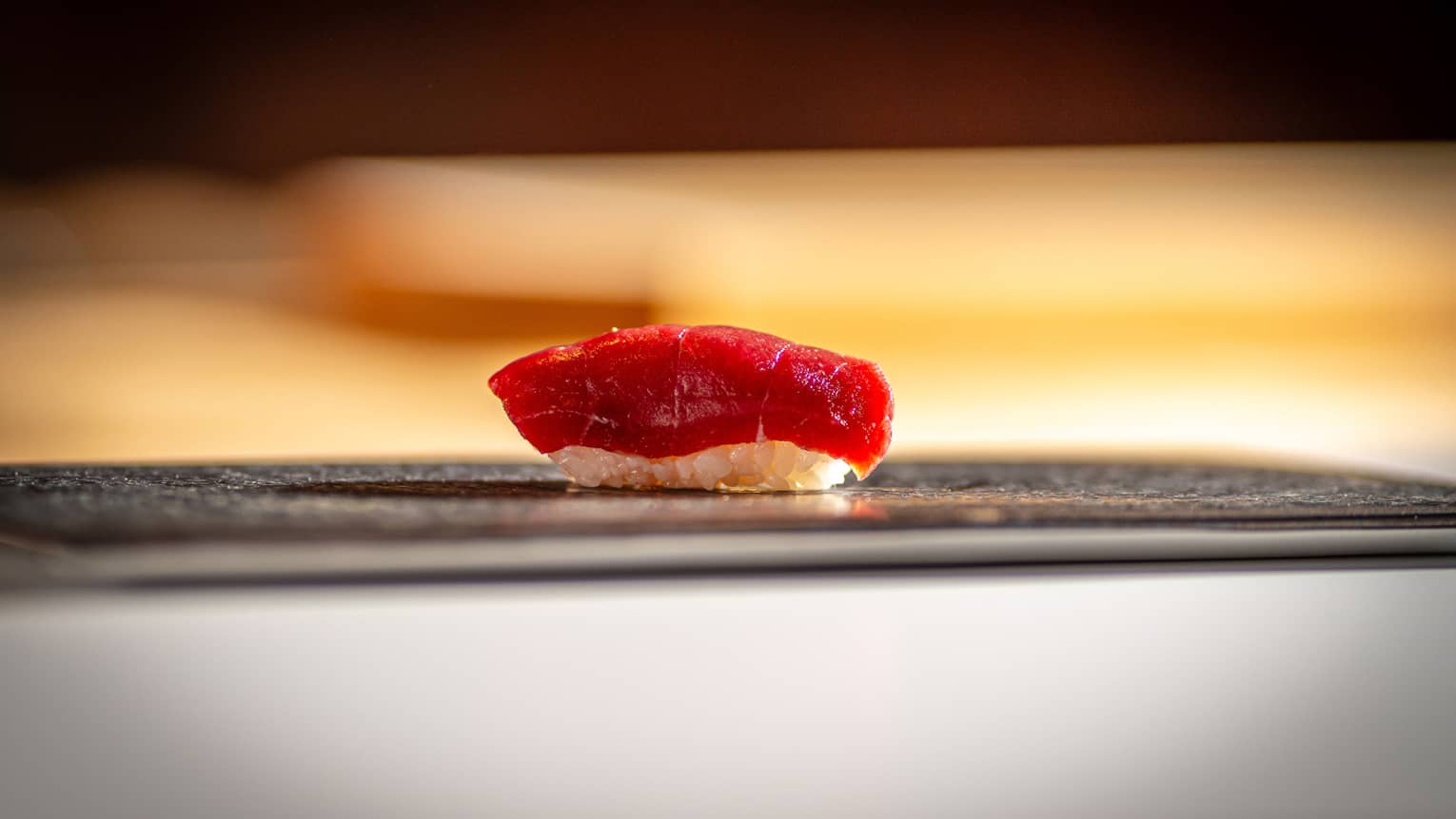 A piece of tuna nigiri sushi displayed on a thin dark platter. 