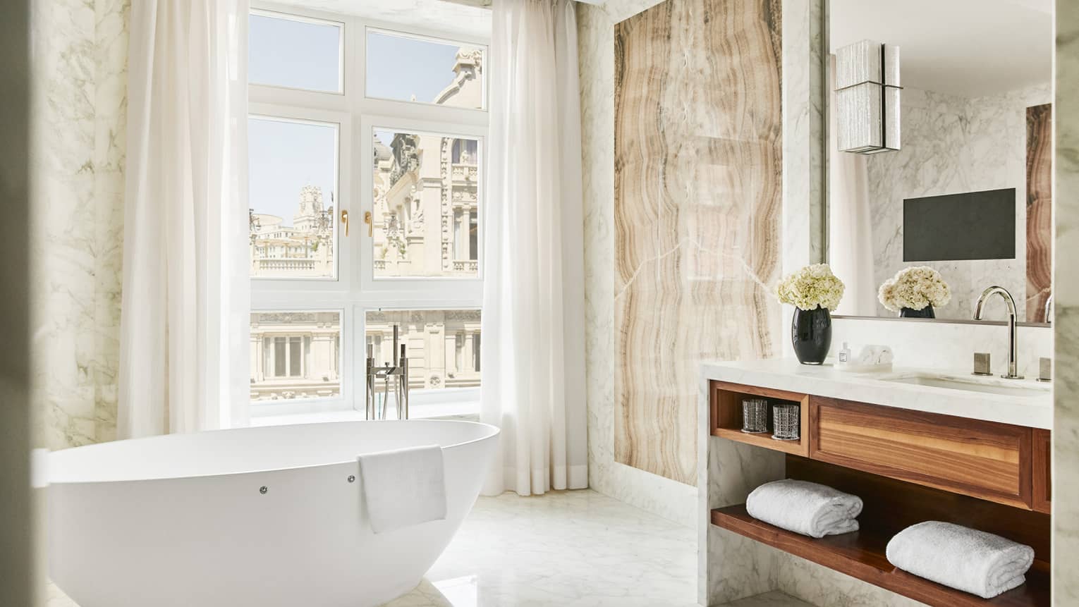 Luxury hotel bathroom with a freestanding bathtub, marble walls, vanity with wooden shelving, mirror, TV, large window with city view and elegant decor