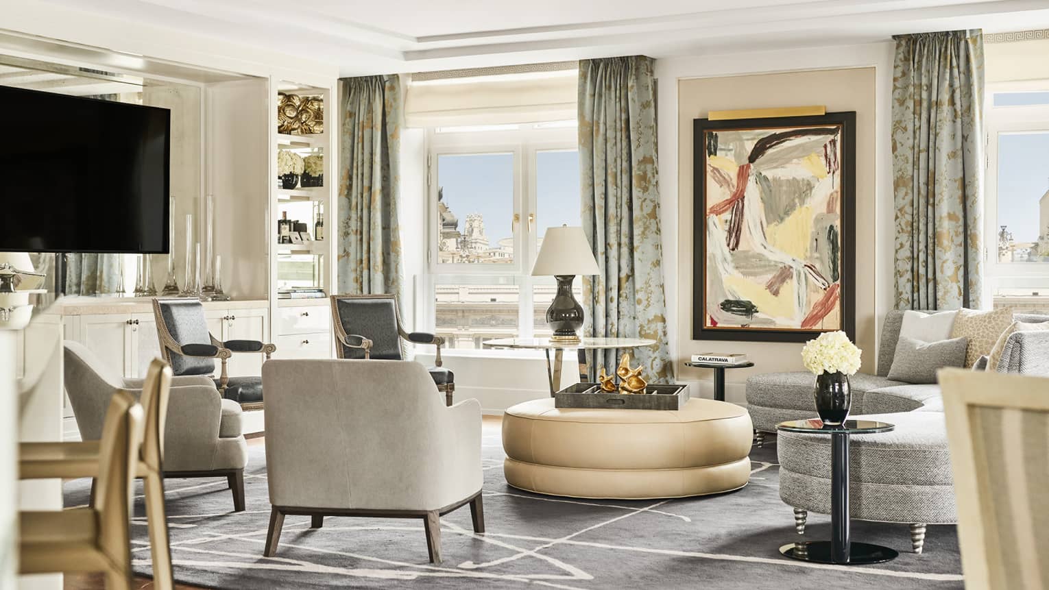 Hotel suite with abstract artwork, TV, grey armchairs and sofa, round beige ottoman, modern decor, glass tables and floor-to-ceiling windows with city views