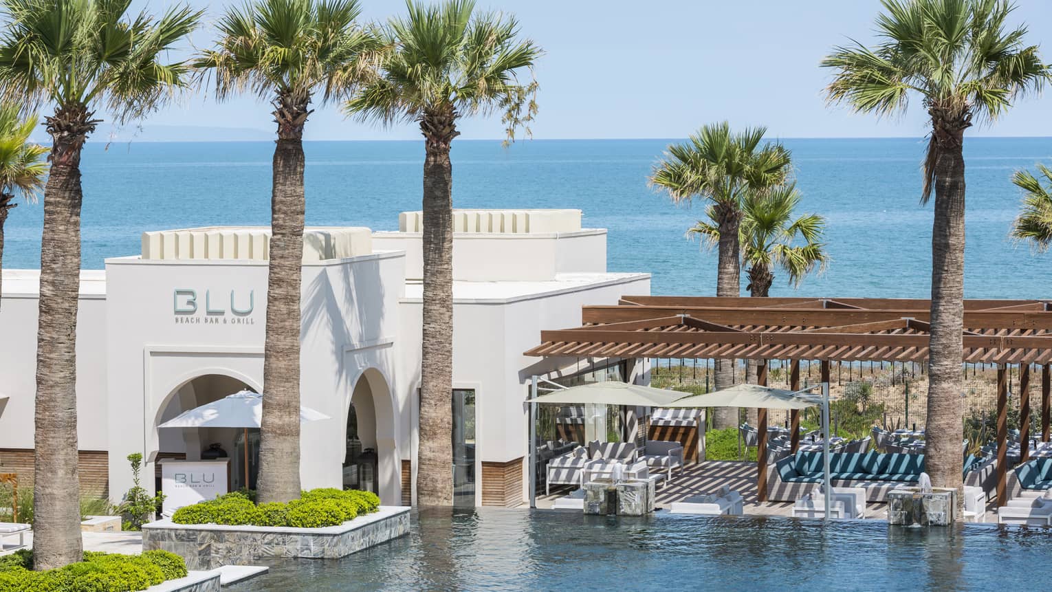 Exterior of BLU Bar & Grill’s white building beside pool surrounded by palm trees, sea in backdrop