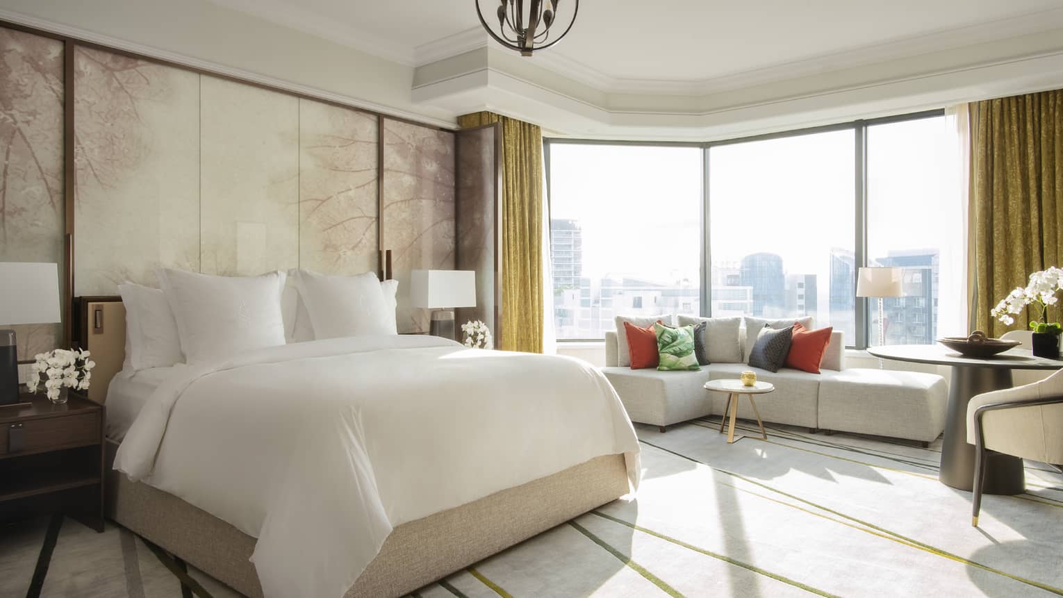 A spacious guest room featuring a large bed, seating area with colourful pillows and floor-to-ceiling windows with city views.