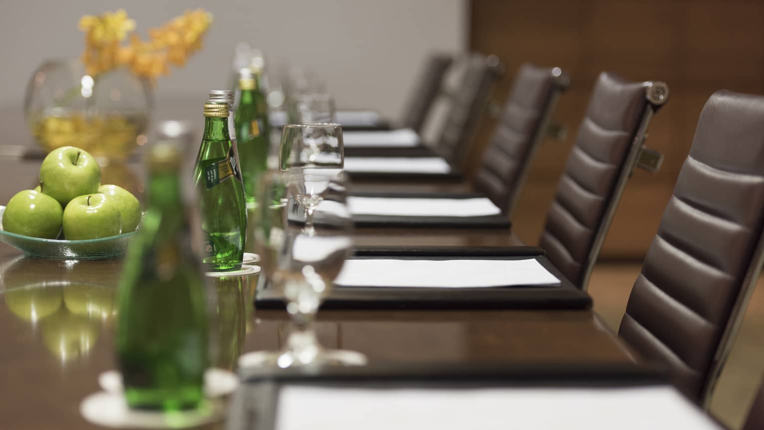 Close-up of business meeting table row of chairs, paper agendas, green glass bottles, apples