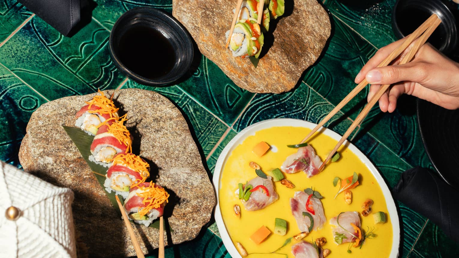 Three people use chopsticks to take colourful food items from decorative serving plates set on a green tiled tabletop.