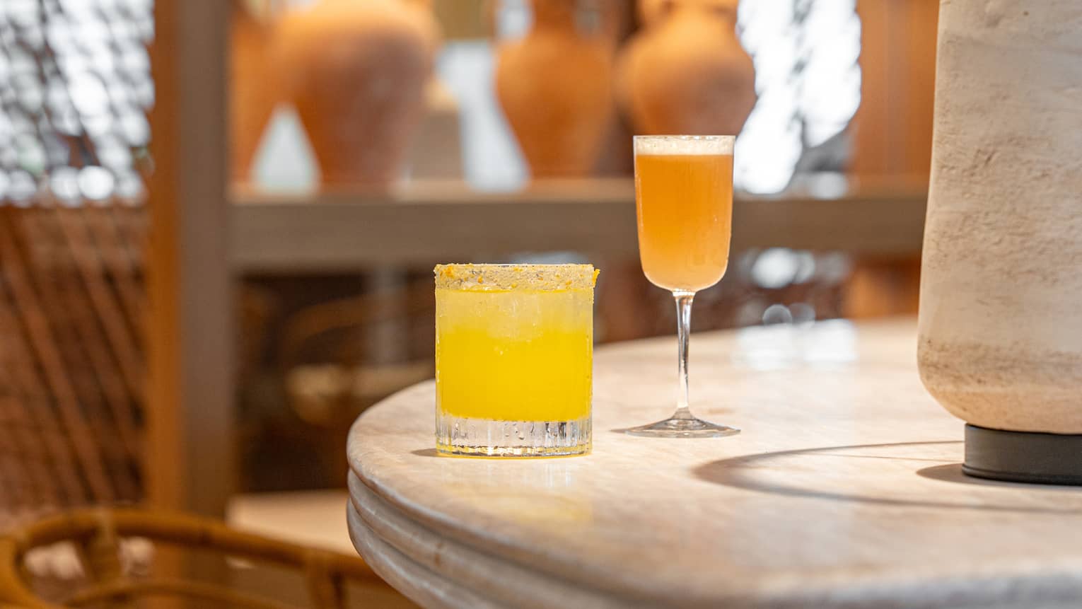 A bright yellow cocktail in a tumbler and a golden cocktail in a stemmed glass sit next to a lamp on a table in an indoor space. Chairs are next to the table, and shelves of vases are in the background.