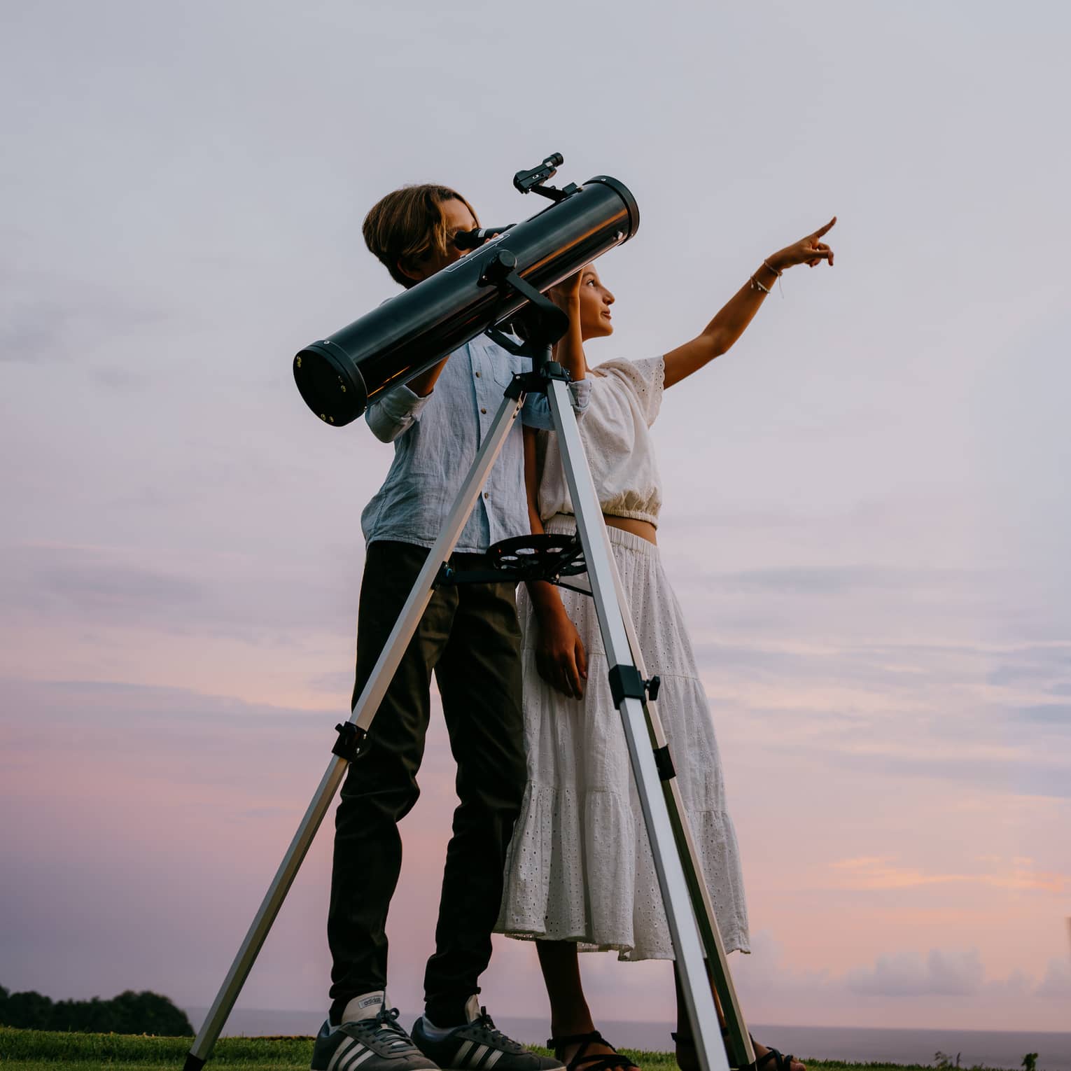 Two people stand next to a large telescope with an evening sky in the background. One person points toward the sky.